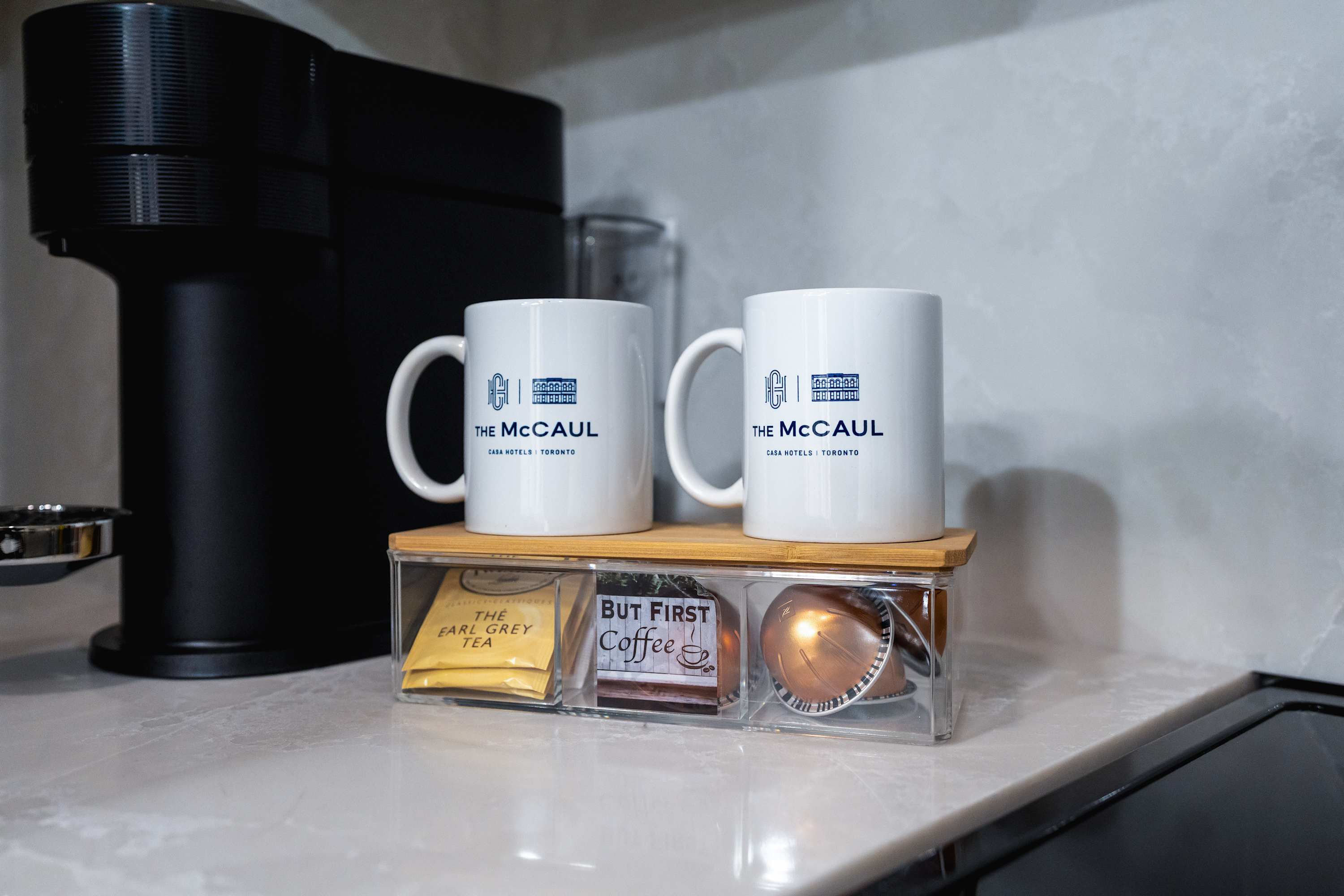 Close-up of a coffee station on a white countertop, featuring a black coffee machine, two white mugs with "The McCaul" logo, and a clear organizer with tea bags and coffee pods.