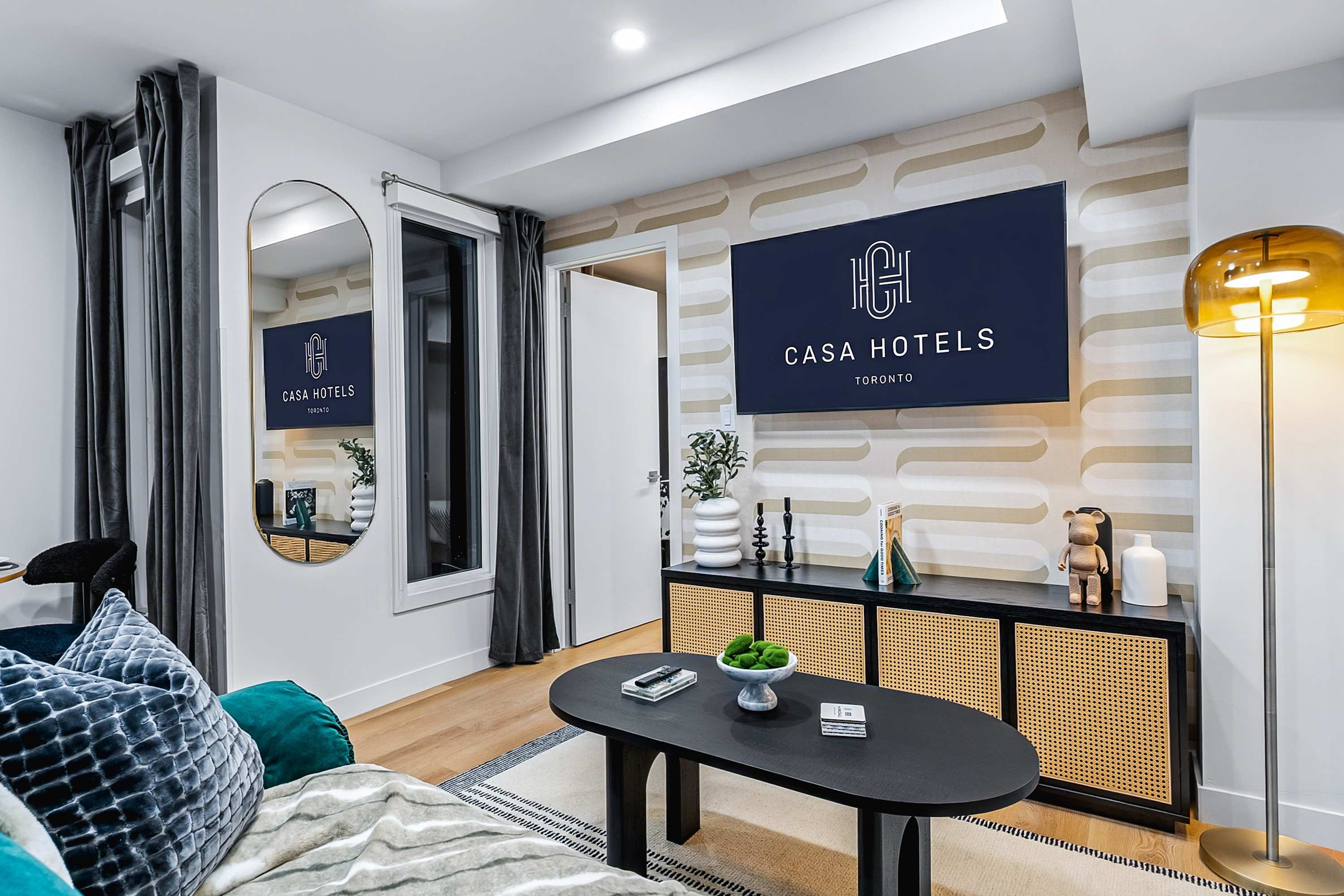 Stylish living room with a dark teal velvet sofa, a black oval coffee table, and a media console with rattan doors under a large TV displaying "CASA HOTELS". A large mirror reflects the room, and an accent wall with a geometric pattern adds visual interest.