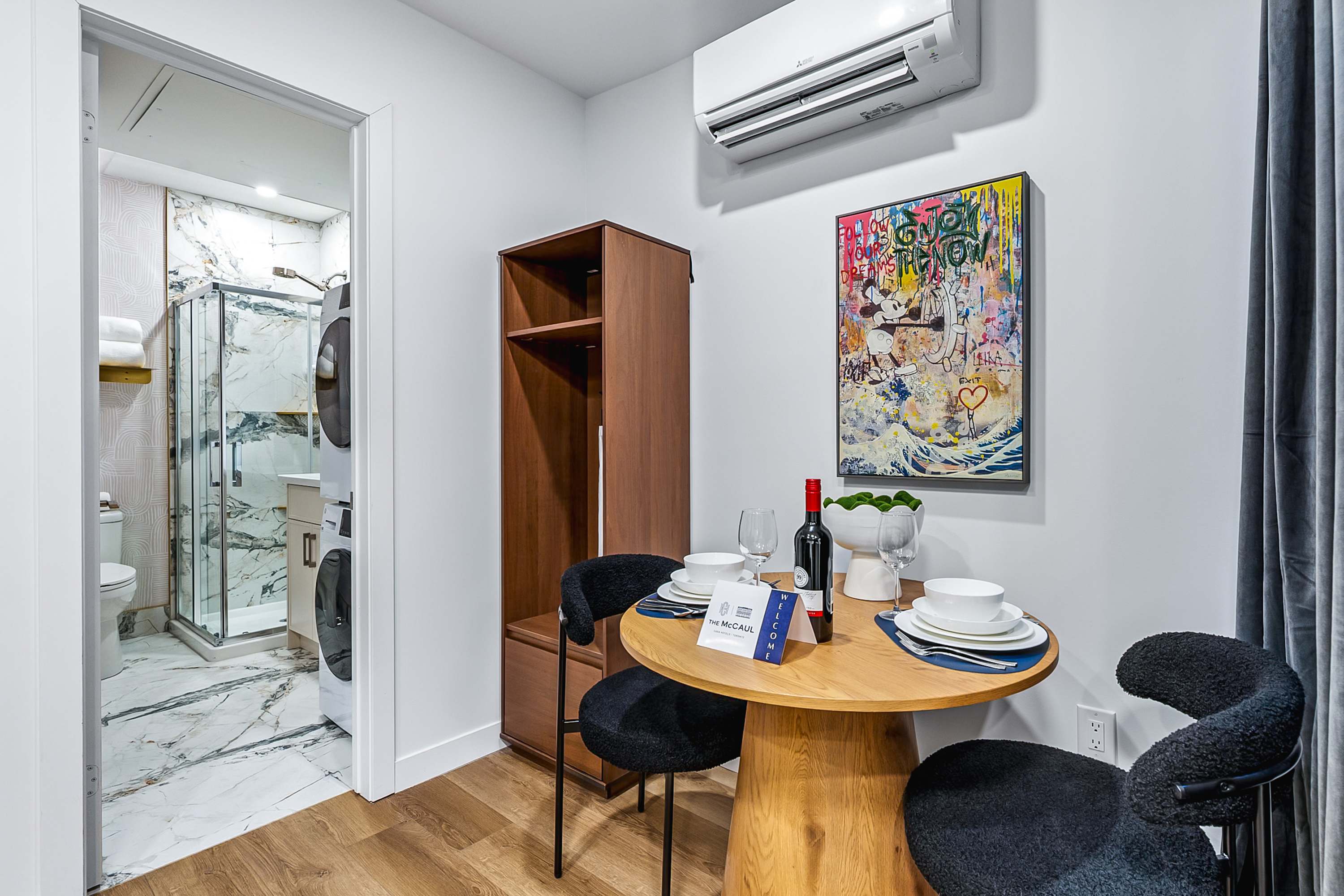 Dining area with a round wooden table and two black textured chairs, next to a wooden storage unit. An open doorway reveals a bathroom with a stacked washer and dryer, and a shower. A colorful abstract painting hangs on the wall.