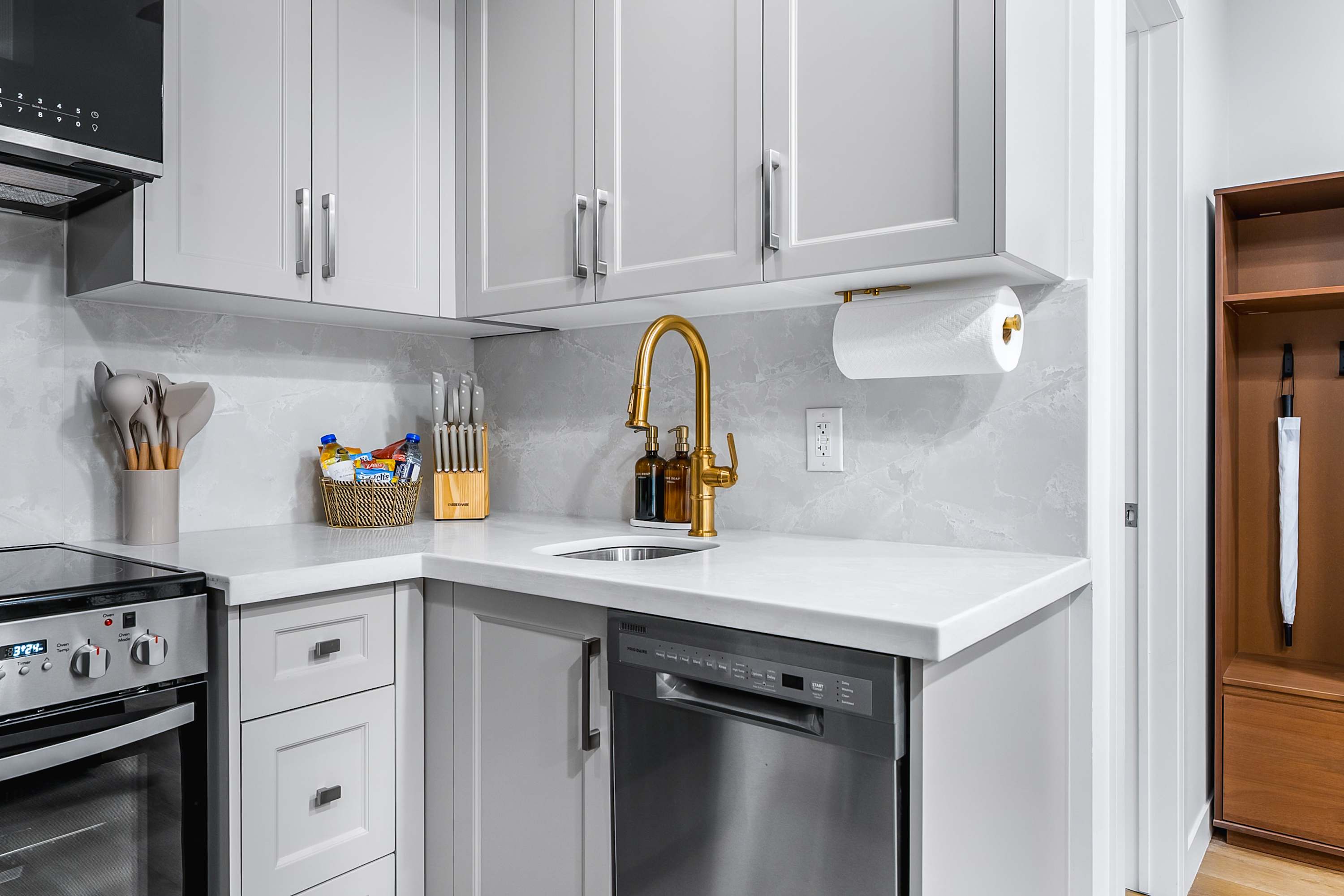 Modern kitchen featuring light gray shaker cabinets, white quartz countertops, a gold-toned faucet, and a gray marble-patterned backsplash. Stainless steel appliances include a dishwasher and oven, with a basket of snacks on the counter.