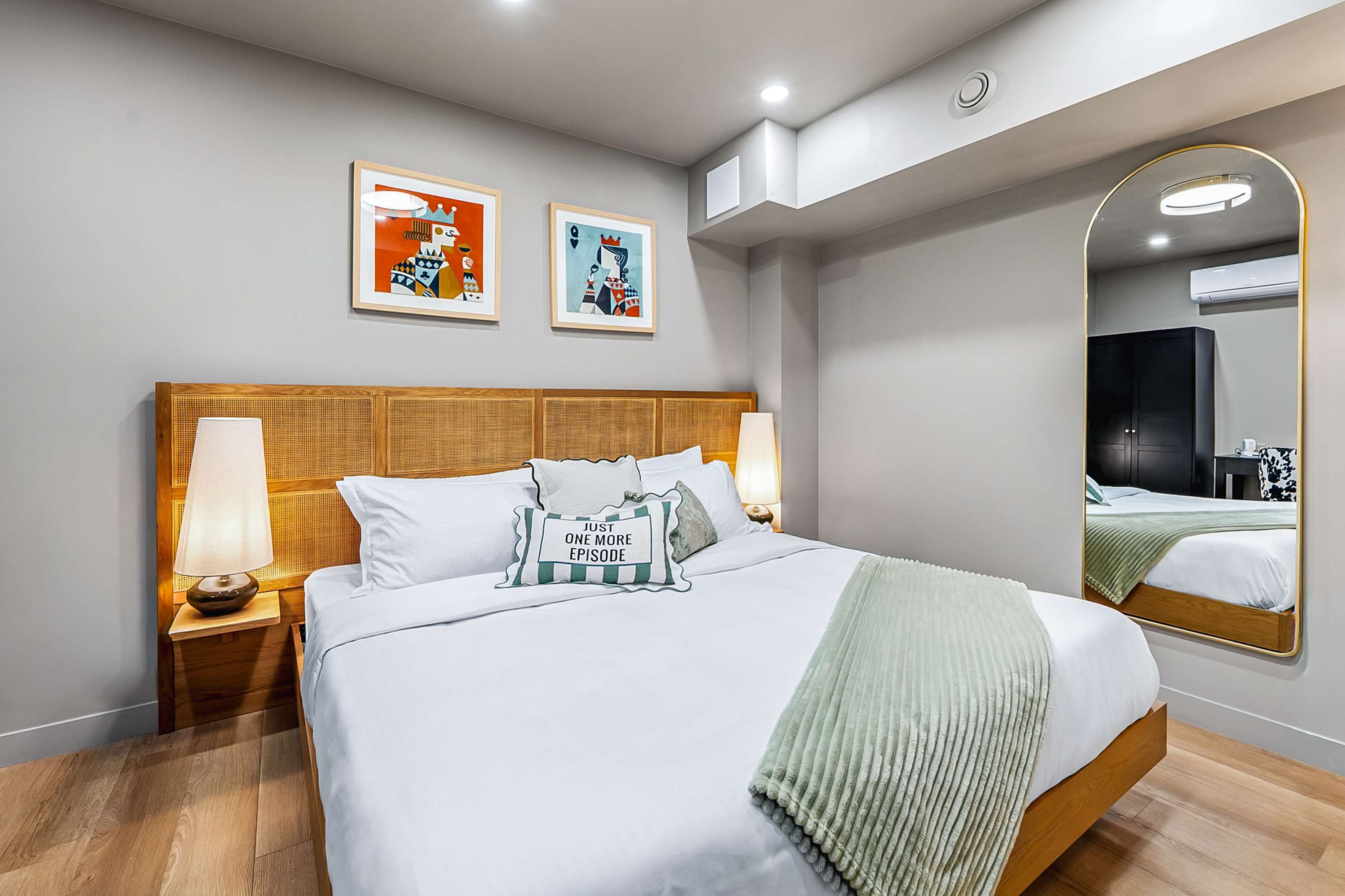Cozy bedroom featuring a queen-sized bed with a woven wooden headboard, white bedding, and a light green throw blanket. Two tall bedside lamps flank the bed, and a large arched gold-framed mirror reflects the room.