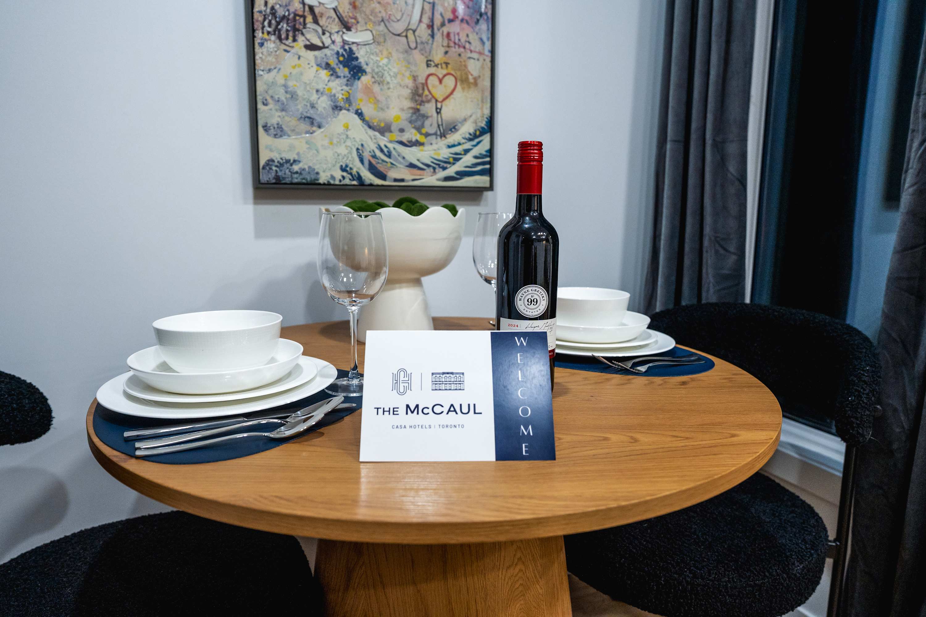 Round wooden dining table set for two with white dinnerware, blue placemats, wine glasses, and a bottle of red wine. A "Welcome" card from "The McCaul" is prominently displayed.