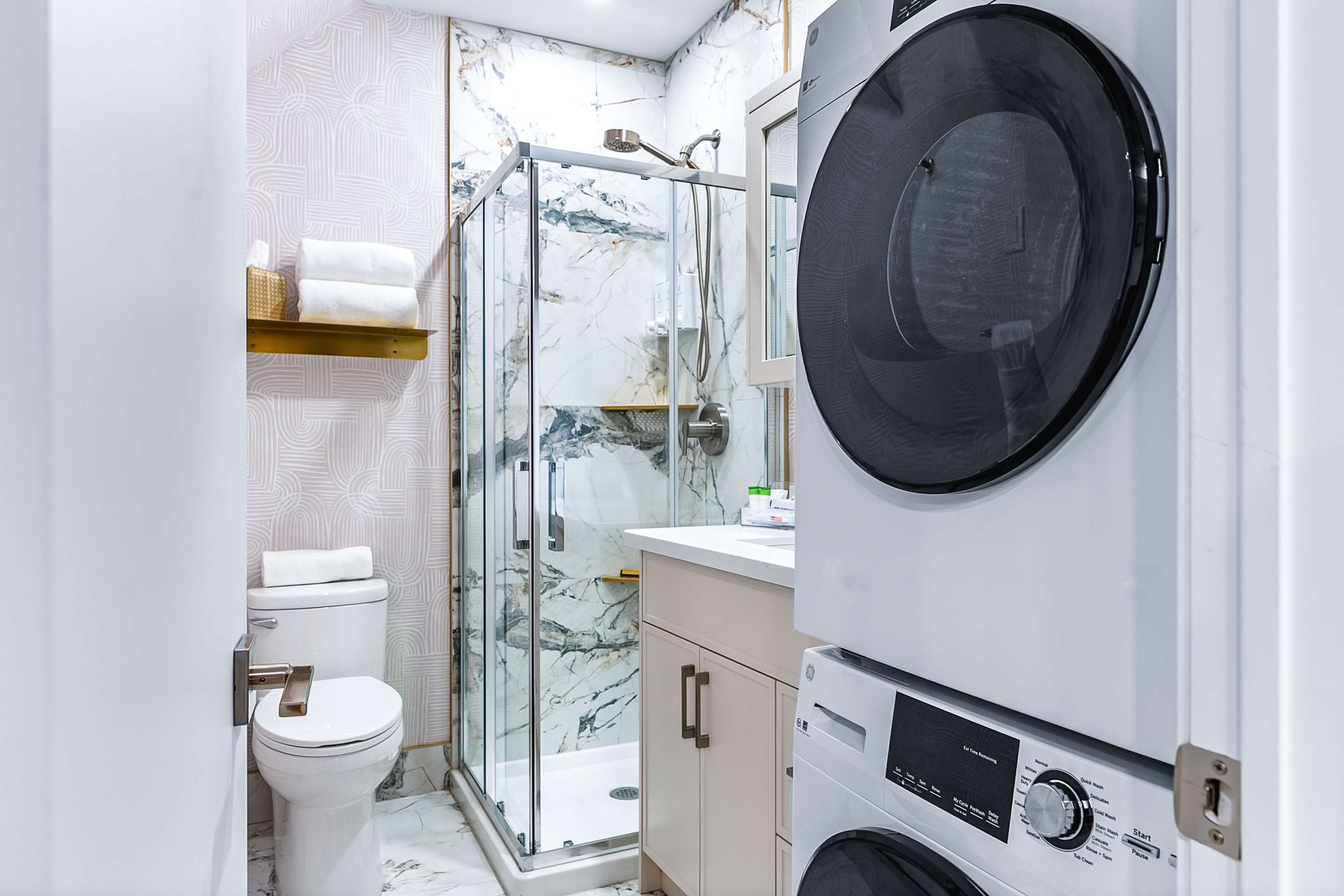 Compact bathroom with a toilet, glass-enclosed shower with marble-patterned tiles, and a stacked washer and dryer unit. A vanity with a sink is visible, and white towels are stacked on a shelf.