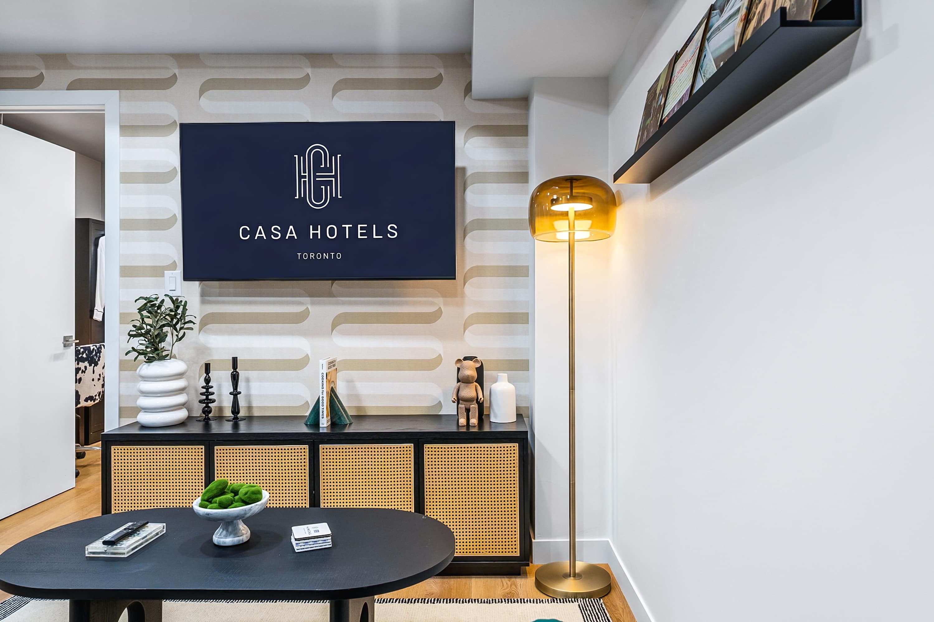 Close-up of a modern living room area, highlighting a black media console with rattan doors, decorative vases, and a large TV displaying "CASA HOTELS". A warm amber floor lamp stands beside the console, illuminating the patterned accent wall.
