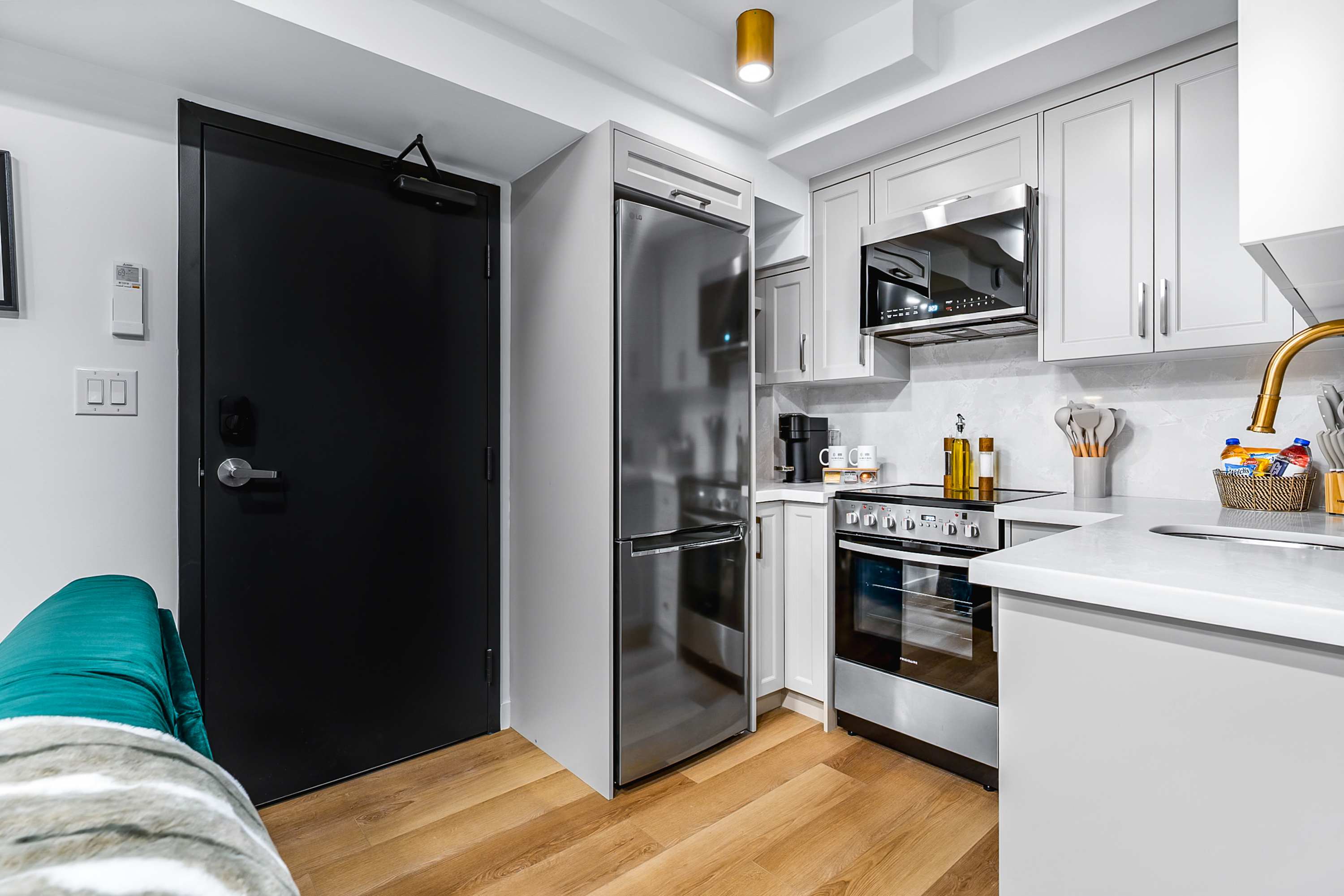 View of the compact kitchen from the living area, showing light gray shaker-style cabinets, white countertops, and stainless steel appliances including a refrigerator and microwave. A black entry door is visible to the left.