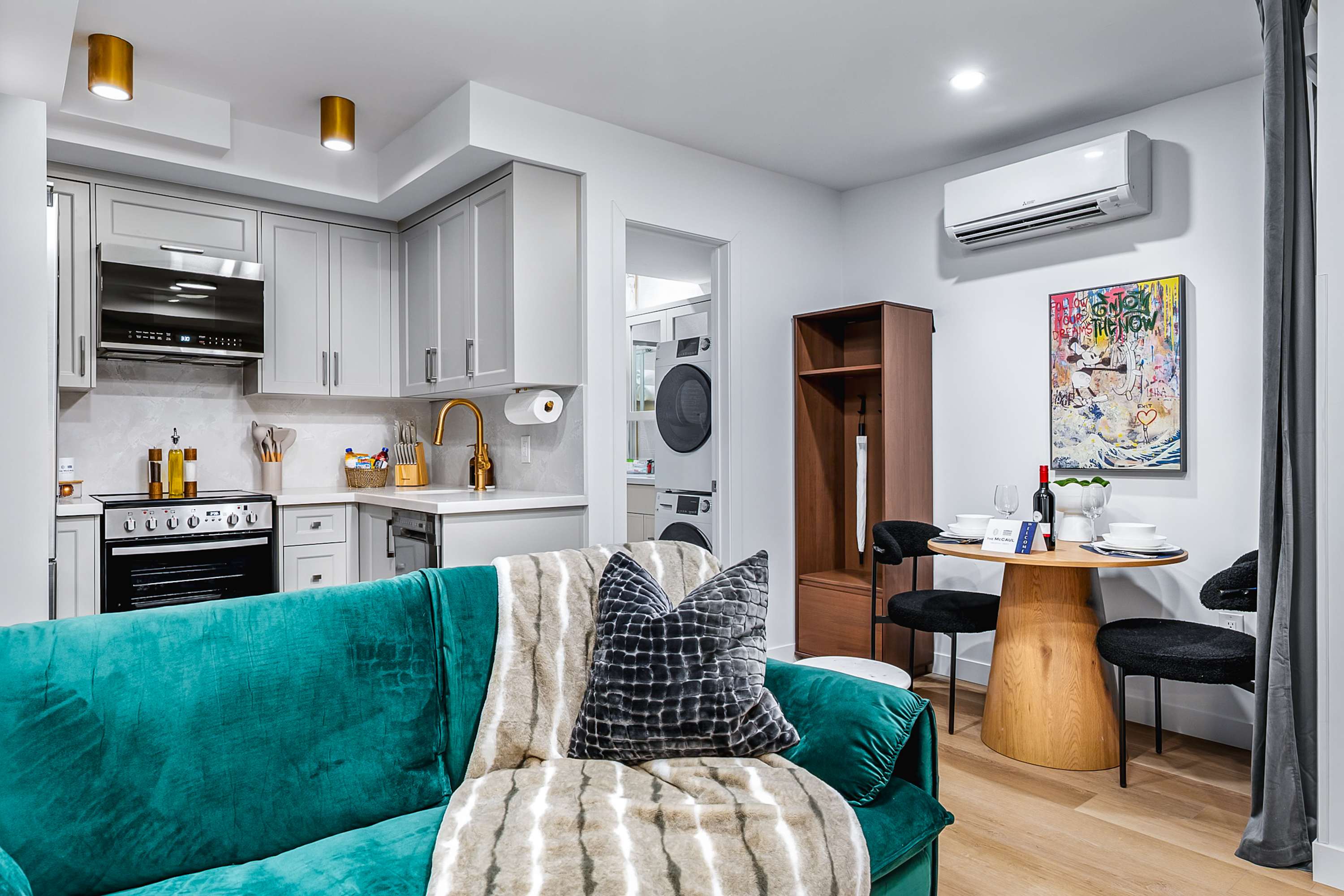 Open-concept living space featuring a teal velvet sofa, a compact kitchen with light gray cabinets, and a dining area with a round wooden table. A glimpse into a laundry area with a washer and dryer is visible through a doorway.