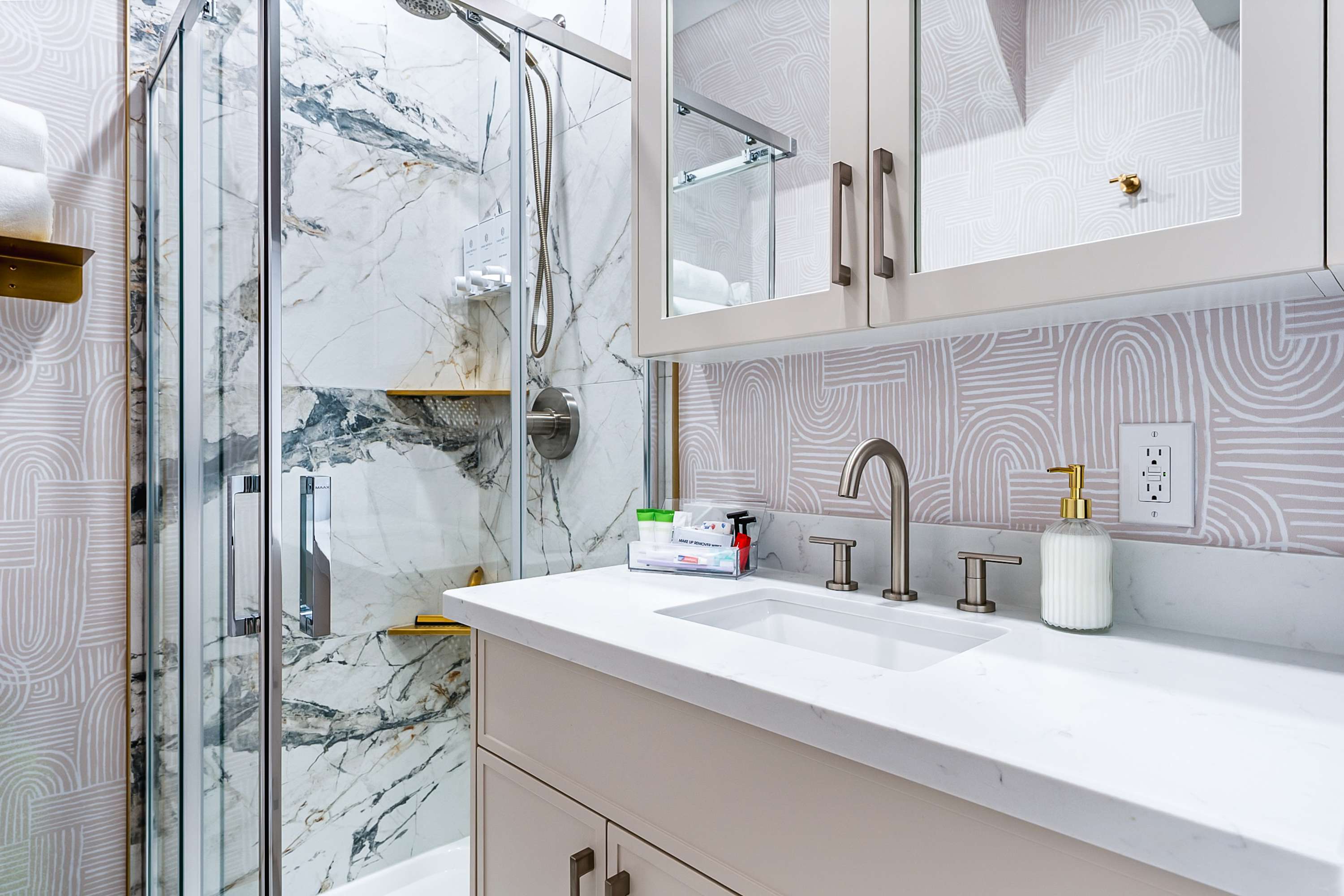 Bathroom featuring a white vanity with a sink and brushed nickel faucet, adjacent to a glass-enclosed shower with marble-patterned tiles. A patterned wallpaper backsplash adds texture to the space.