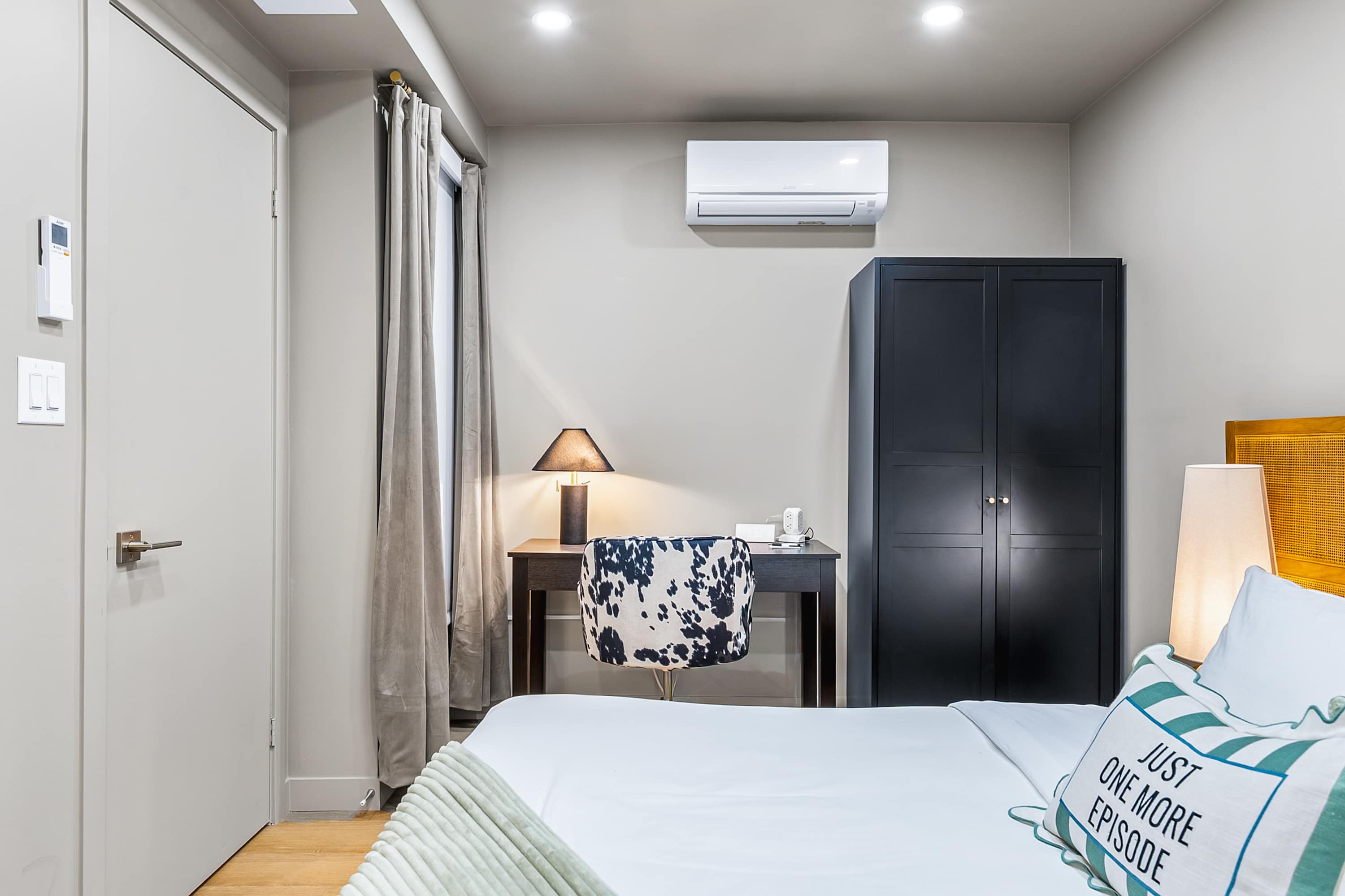 Bedroom view showing a dark wooden desk with a cow-print chair, a black wardrobe, and a window with gray curtains. An air conditioning unit is mounted above the desk.