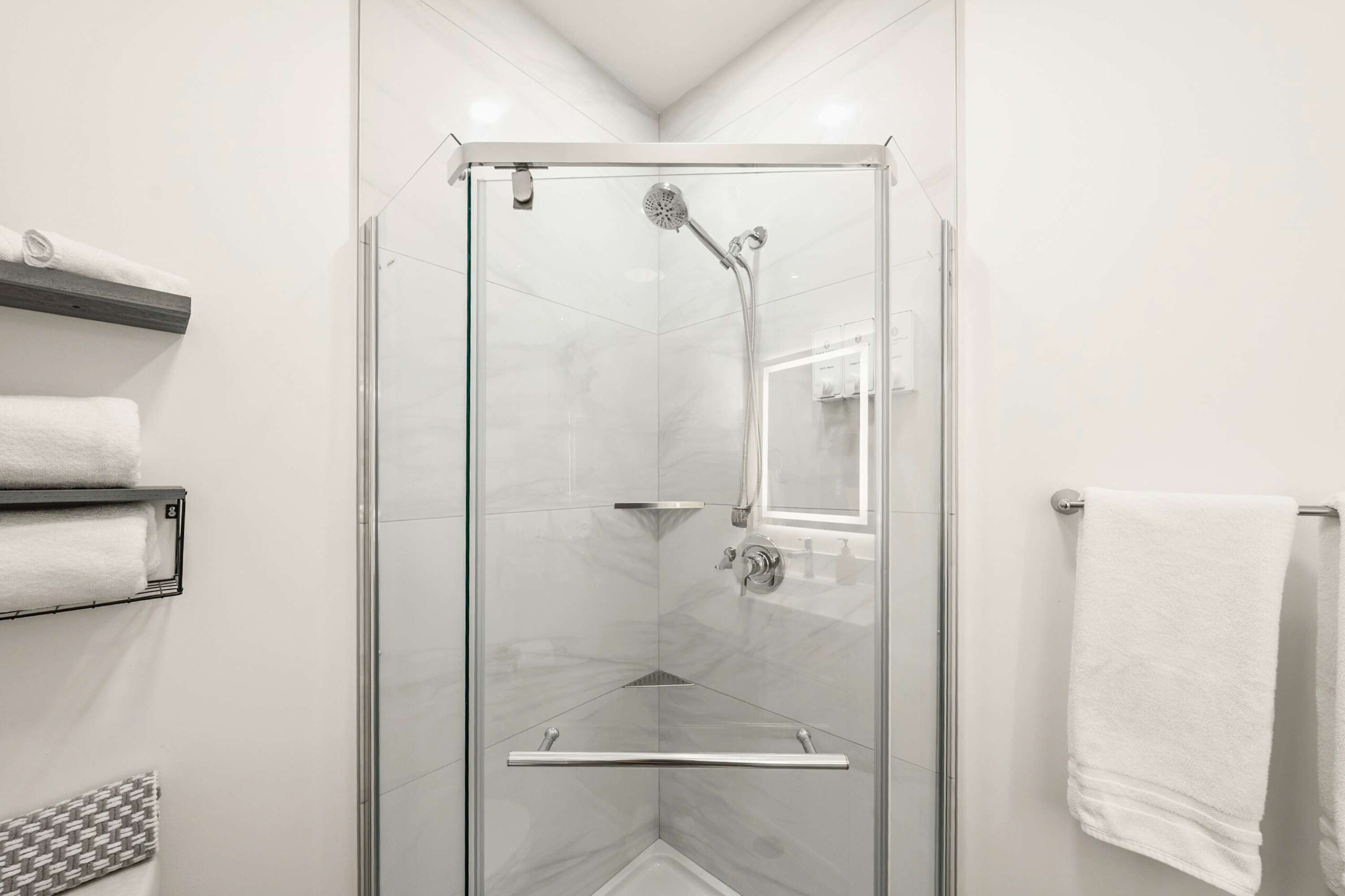 Modern corner shower with clear glass enclosure, white marble-patterned tile walls, and a rain shower head. Two small corner shelves are built into the tile, and white towels are neatly folded on a nearby shelf.