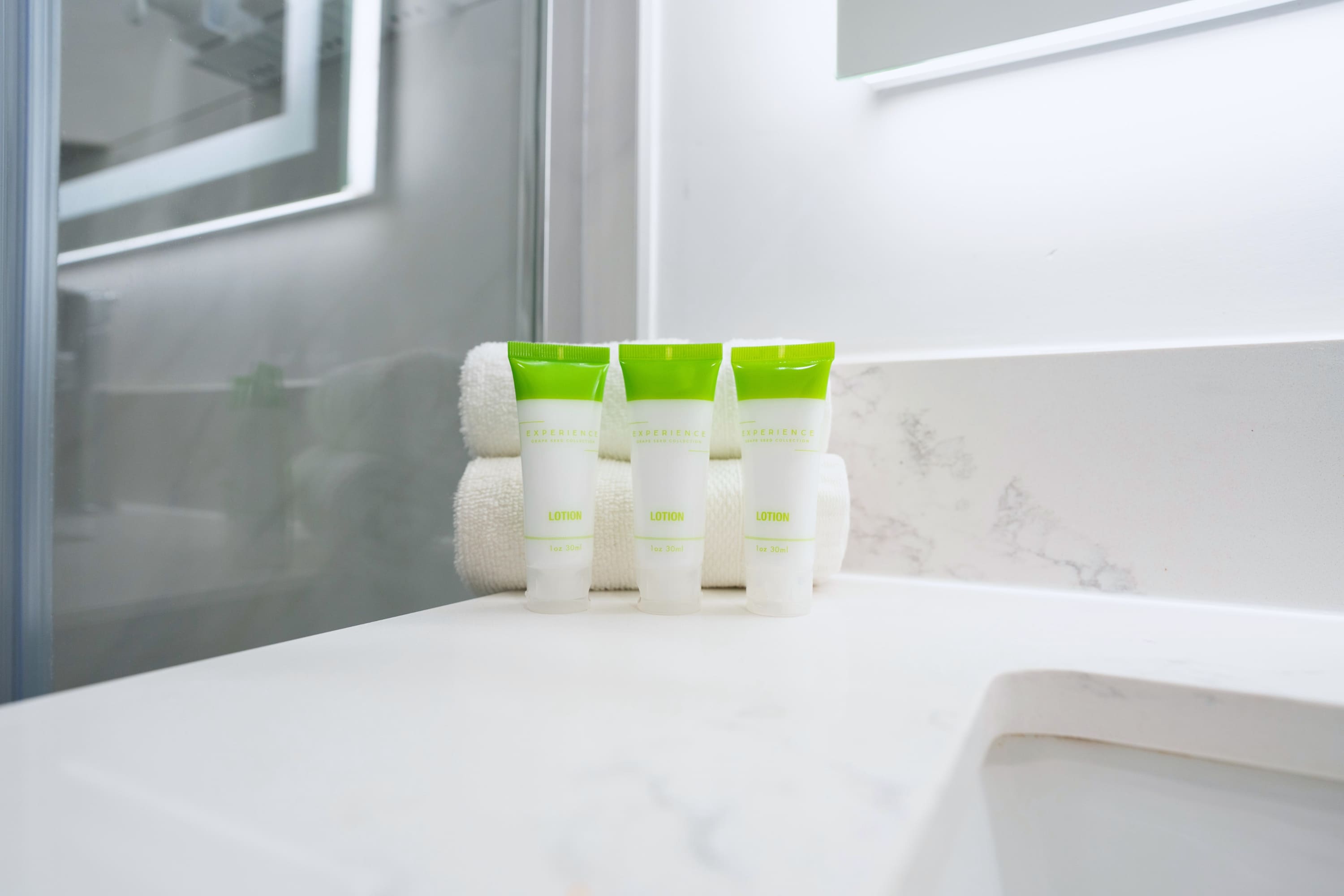 A close-up of three small green-capped lotion tubes neatly arranged on a clean white bathroom counter, with soft white towels stacked behind them, suggesting a well-stocked vanity.