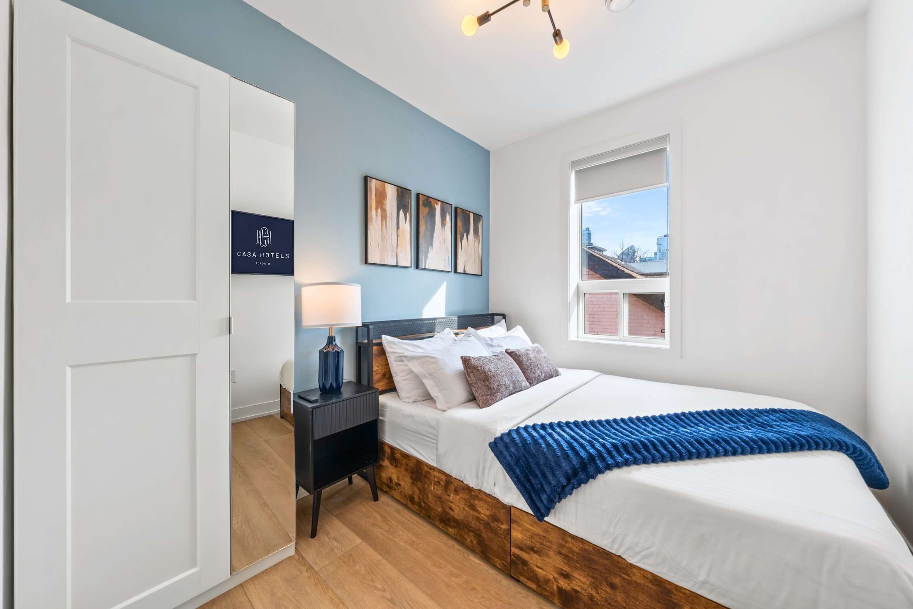 Cozy bedroom featuring a queen-sized bed with a dark headboard, white bedding, and a blue throw blanket, set against a light blue accent wall with abstract artwork. A dark nightstand with a lamp sits beside the bed, and a mirrored closet door reflects a wall-mounted TV.