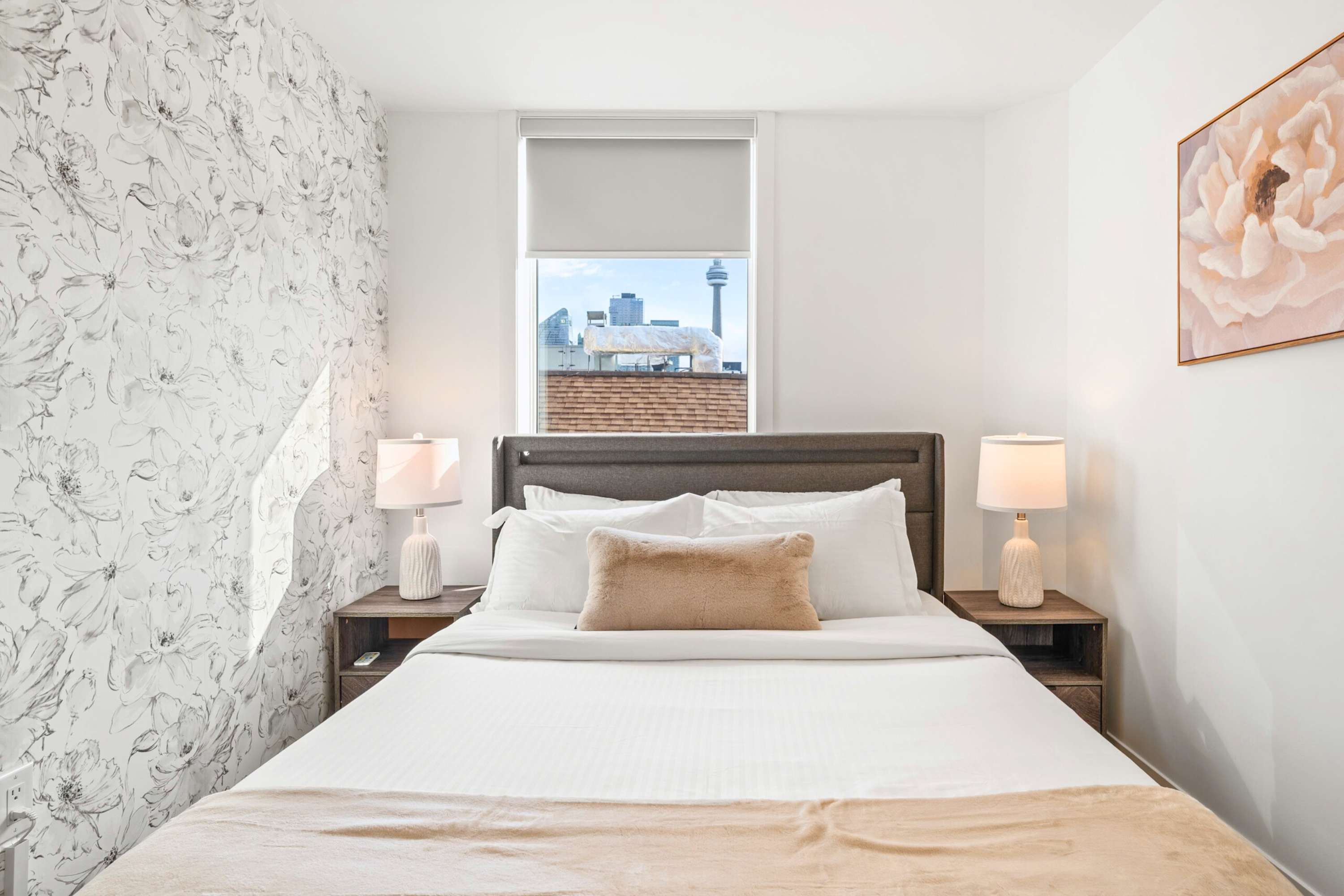 Inviting bedroom with a queen bed, gray headboard, and a floral accent wallpaper on the left wall. A large window offers a city view, including the CN Tower, while two nightstands with lamps provide bedside convenience.