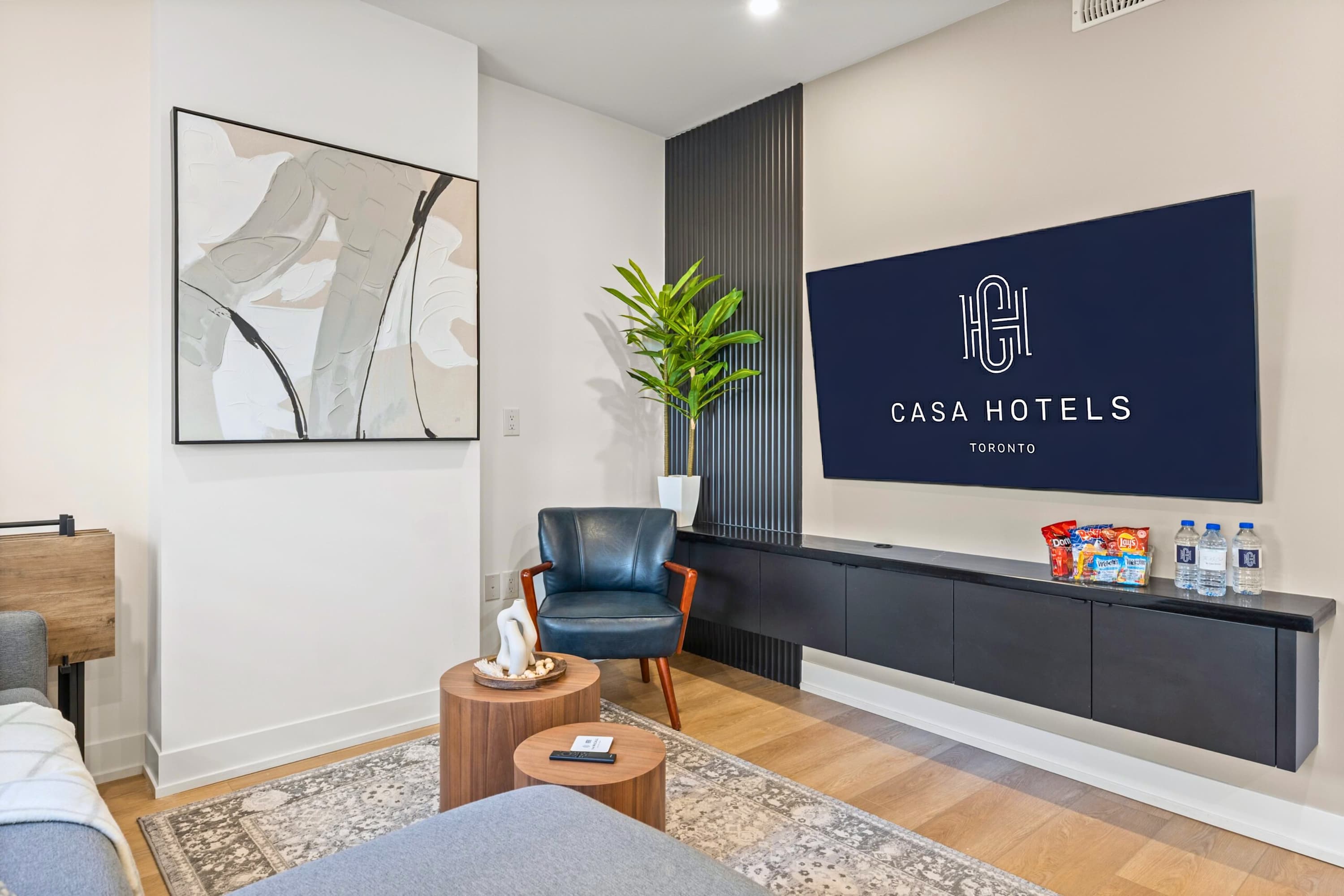 Modern living room with a large wall-mounted TV displaying the 'CASA HOTELS TORONTO' logo, a floating black media console, and a dark slatted accent wall. A dark blue armchair and two round wooden tables sit on a patterned rug.