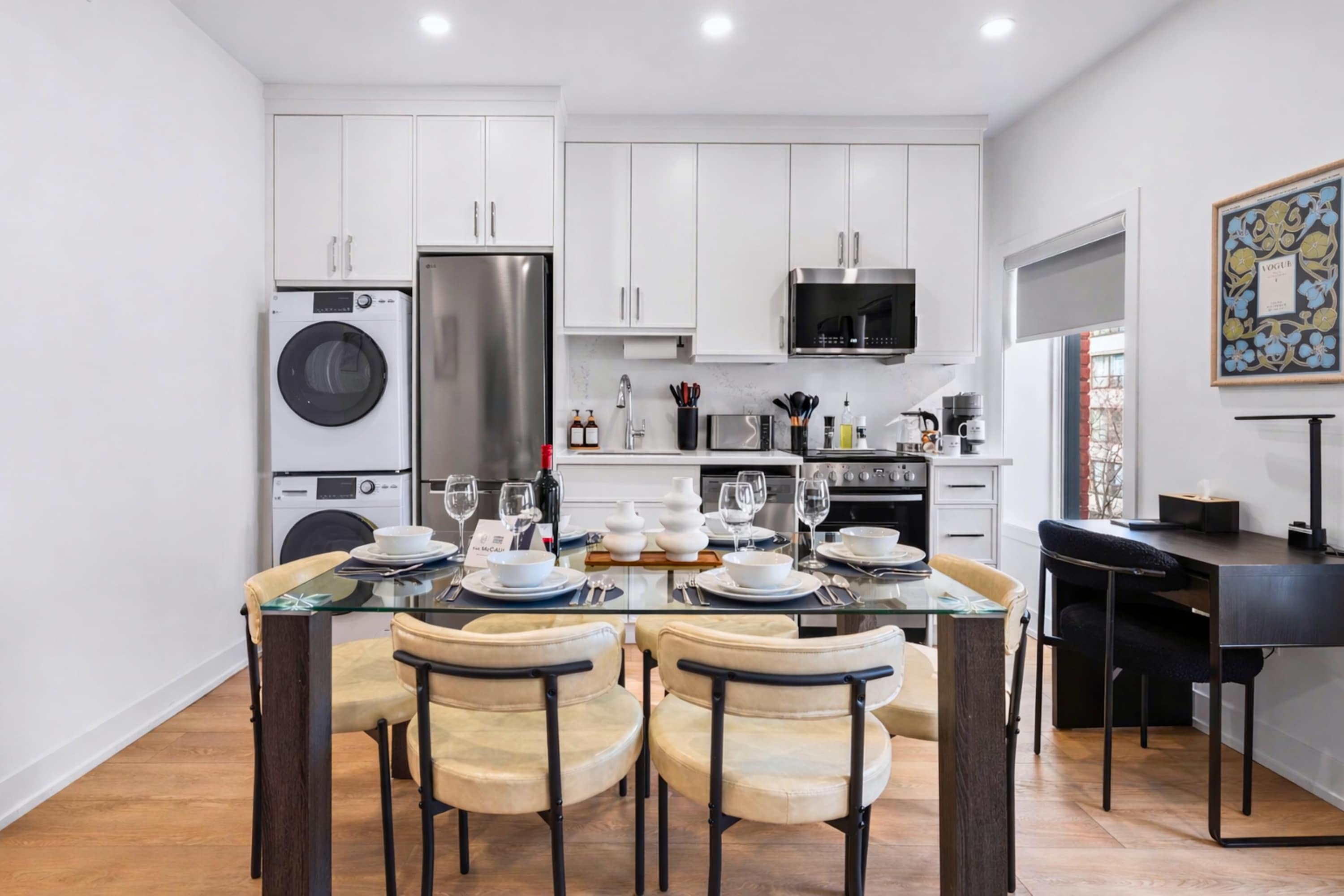 Modern kitchen and dining area featuring white cabinetry, stainless steel appliances including a stacked washer and dryer, and a glass-top dining table set for four. Light wood flooring and a small desk nook are also visible.