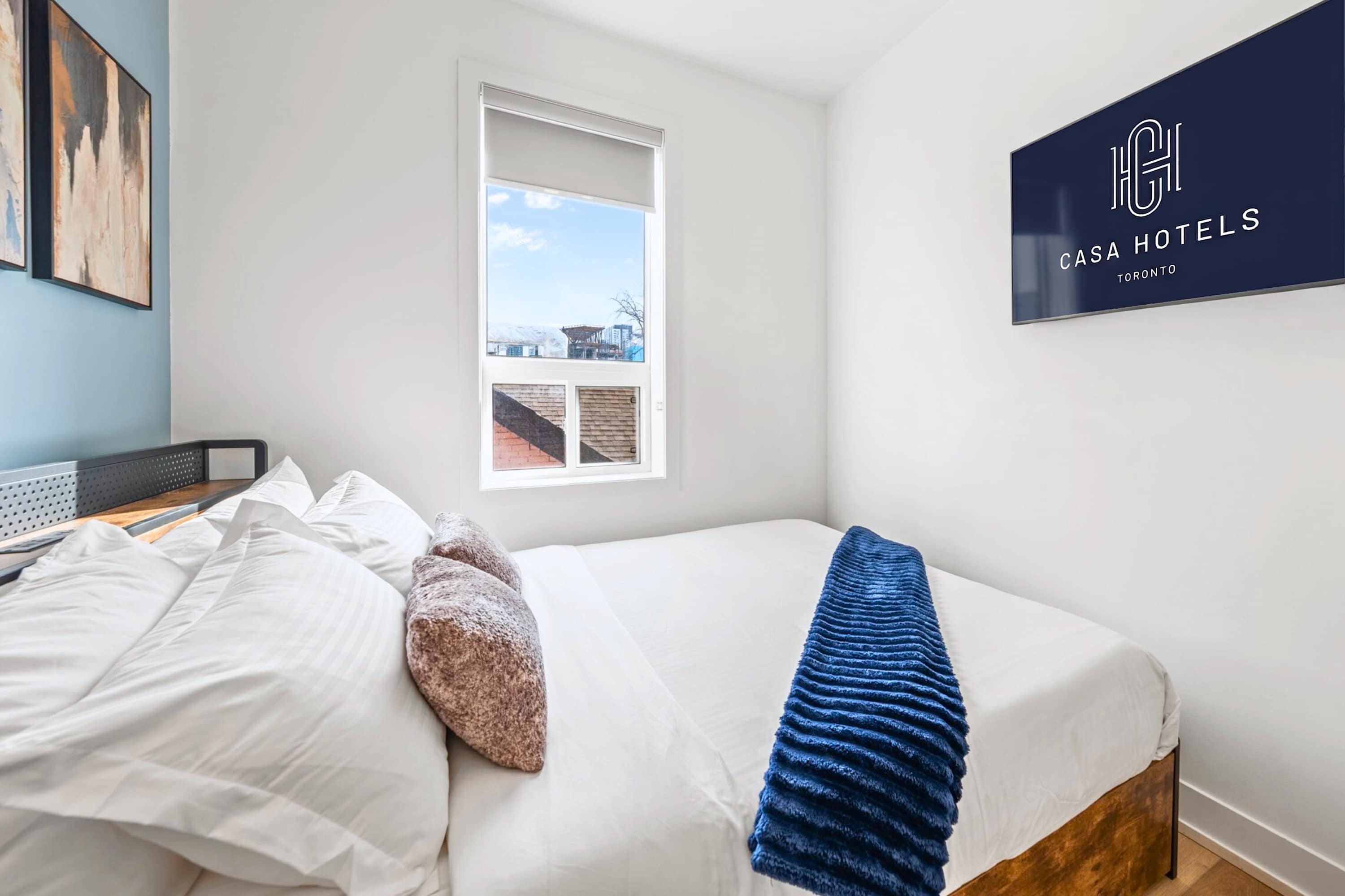 Close-up view of a queen bed with crisp white linens and a textured blue throw blanket, accented by two decorative pillows. A large window with a roller shade offers a glimpse of the city, and a wall-mounted TV displays the 'CASA HOTELS' logo.