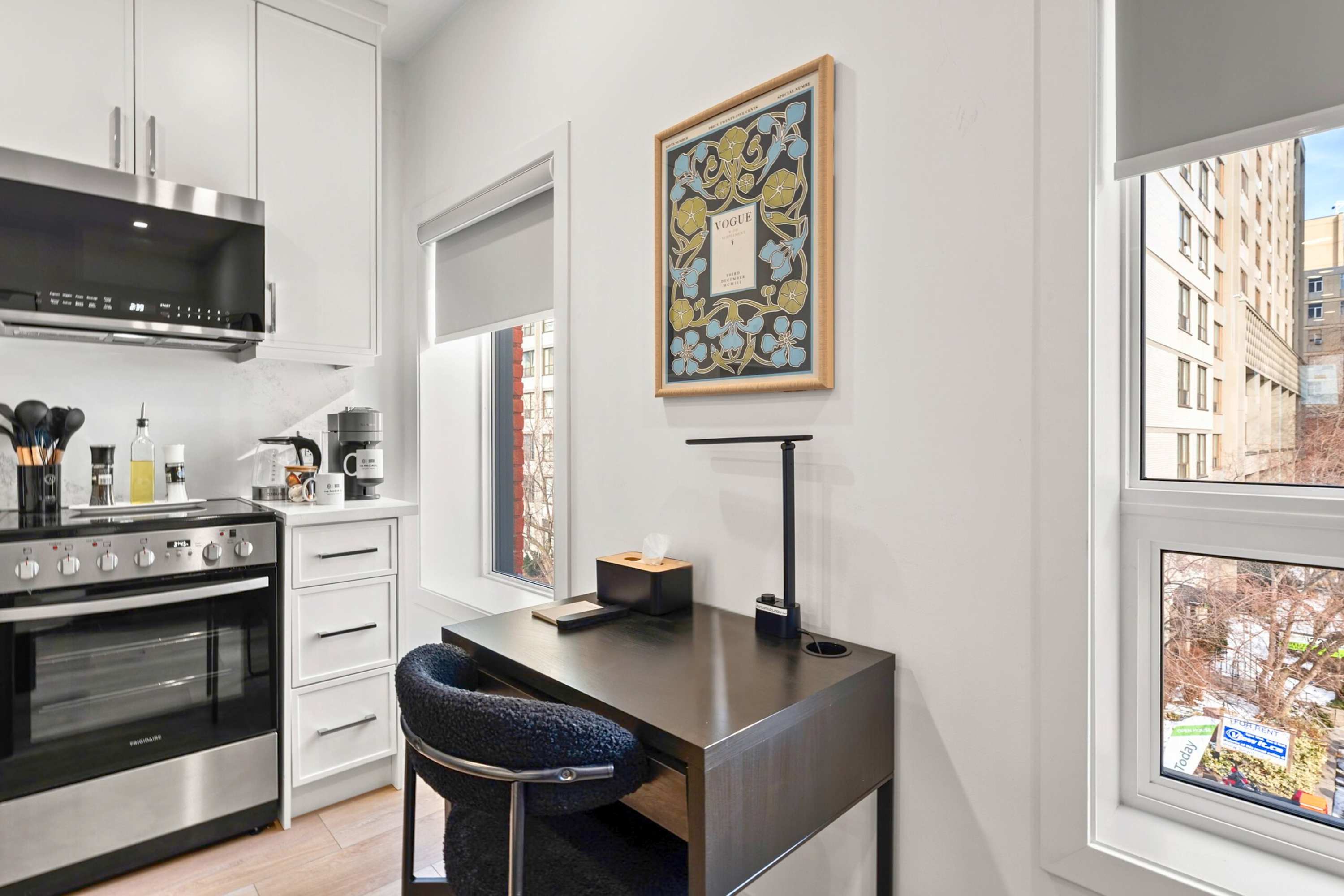 A compact office nook with a dark wooden desk, a black textured chair, and a modern desk lamp, positioned next to a window with a city view. A framed art print hangs on the white wall above.