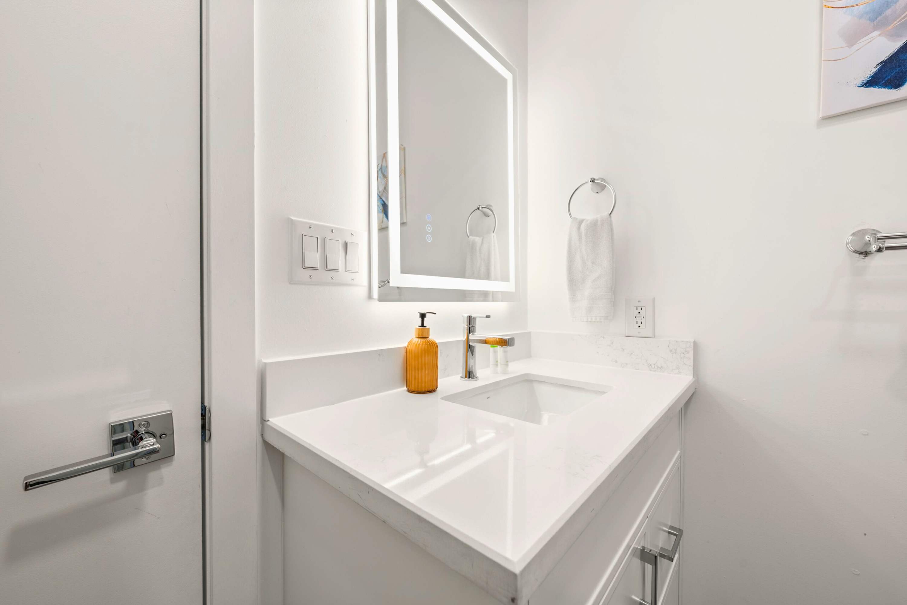 Contemporary bathroom vanity with a white quartz countertop, an integrated sink, and a large LED-backlit mirror. A modern faucet and an orange soap dispenser add a touch of color.