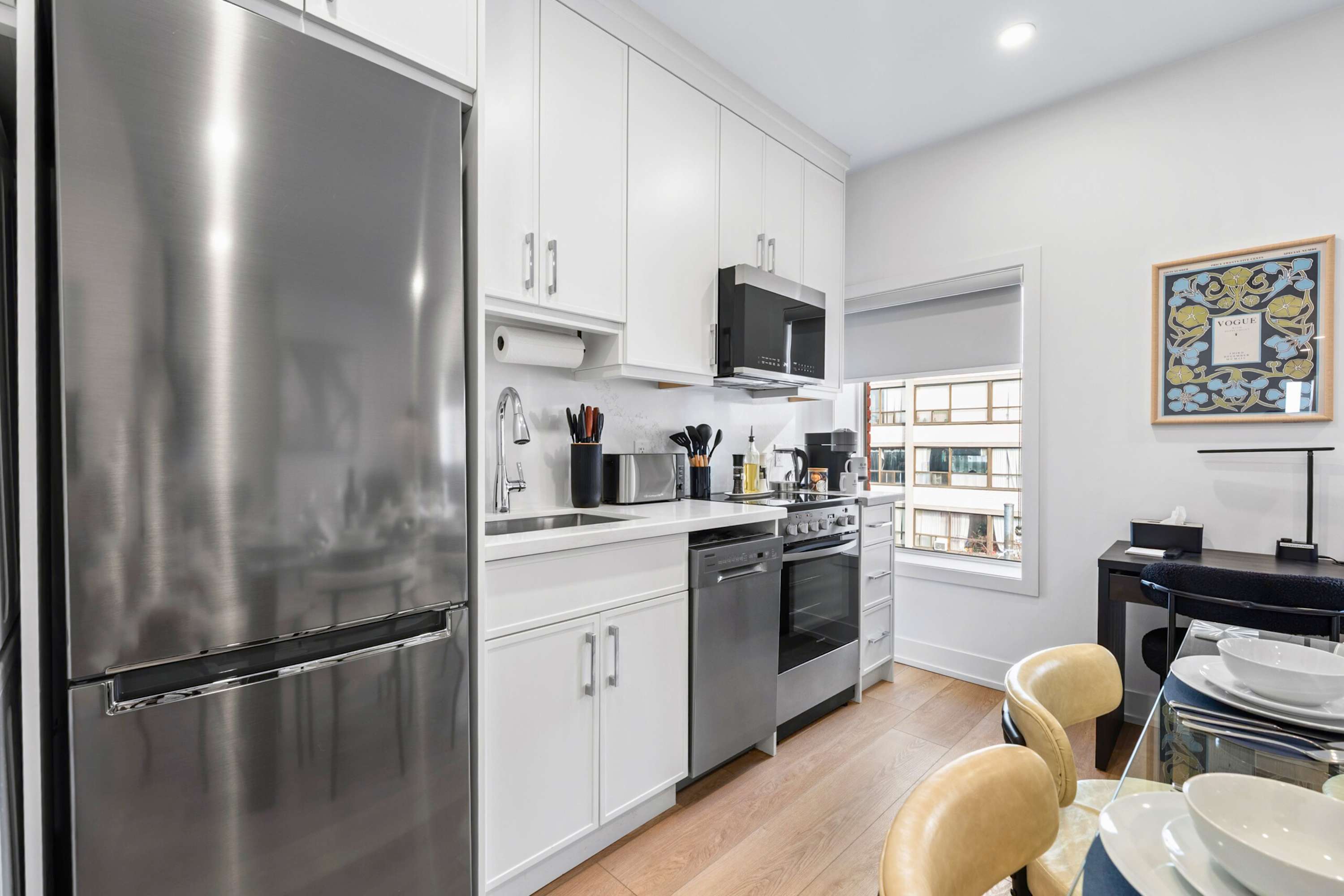 Bright, modern kitchen with white shaker-style cabinets, stainless steel appliances including a large refrigerator, dishwasher, and stove, and light-colored countertops. A dining table is partially visible in the foreground.