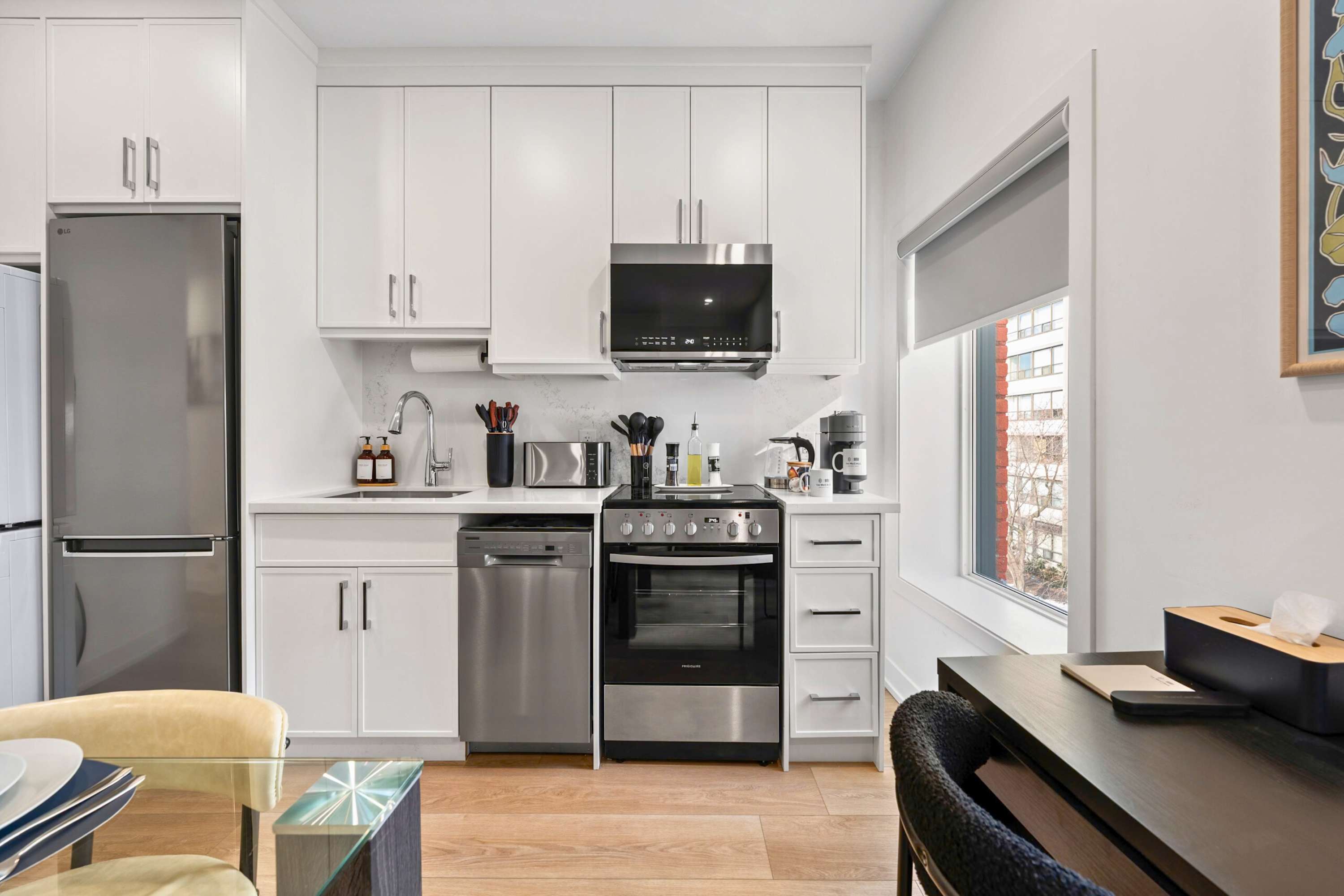 Contemporary kitchen featuring white upper and lower cabinets, stainless steel appliances including a built-in microwave, oven, and dishwasher, and a white quartz countertop. A window provides natural light to the space.