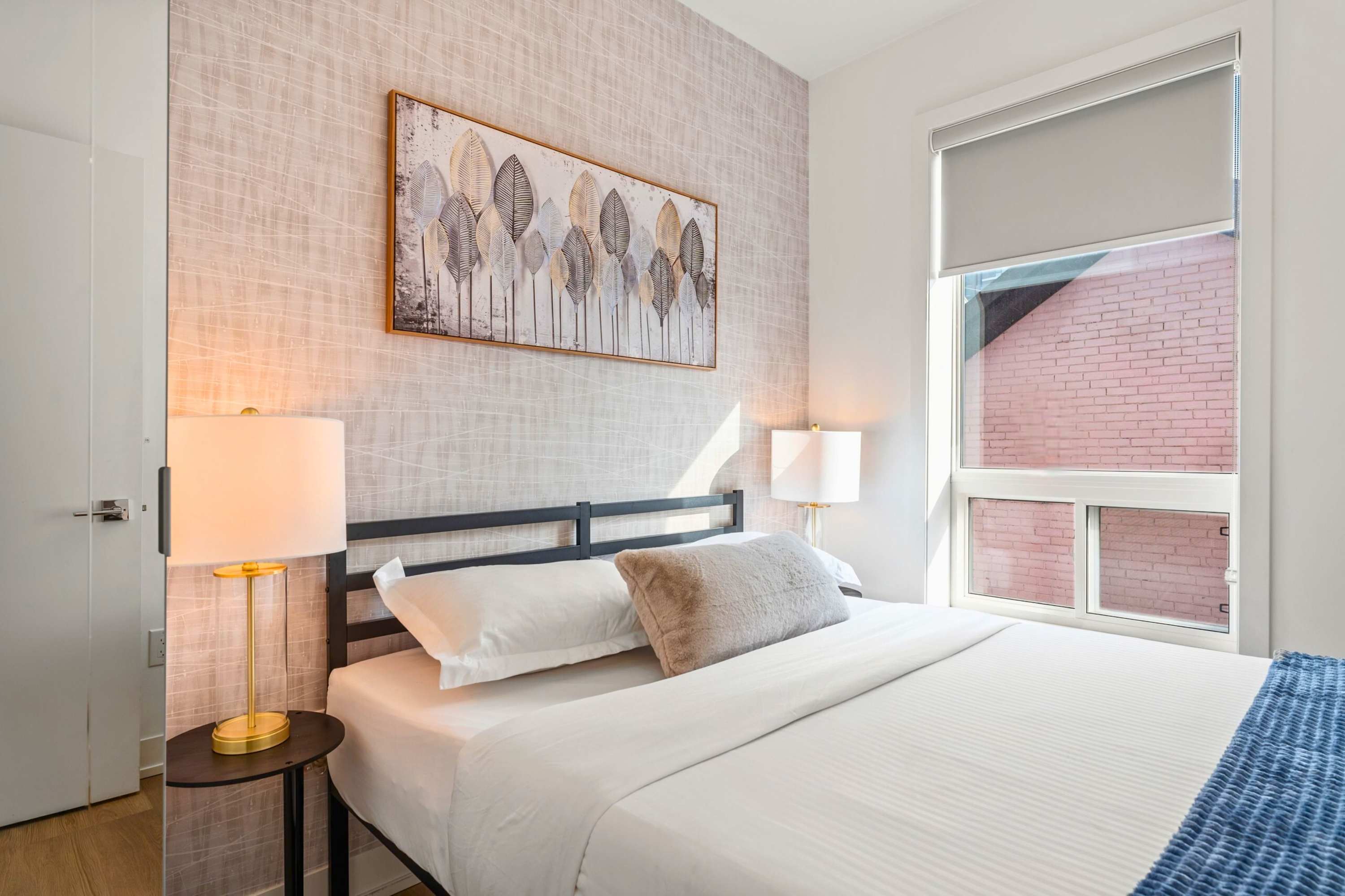 Cozy bedroom featuring a black metal bed frame with white bedding and a grey throw pillow, flanked by two round nightstands with gold-accented lamps. A textured accent wall and a large window with a roller shade are visible.