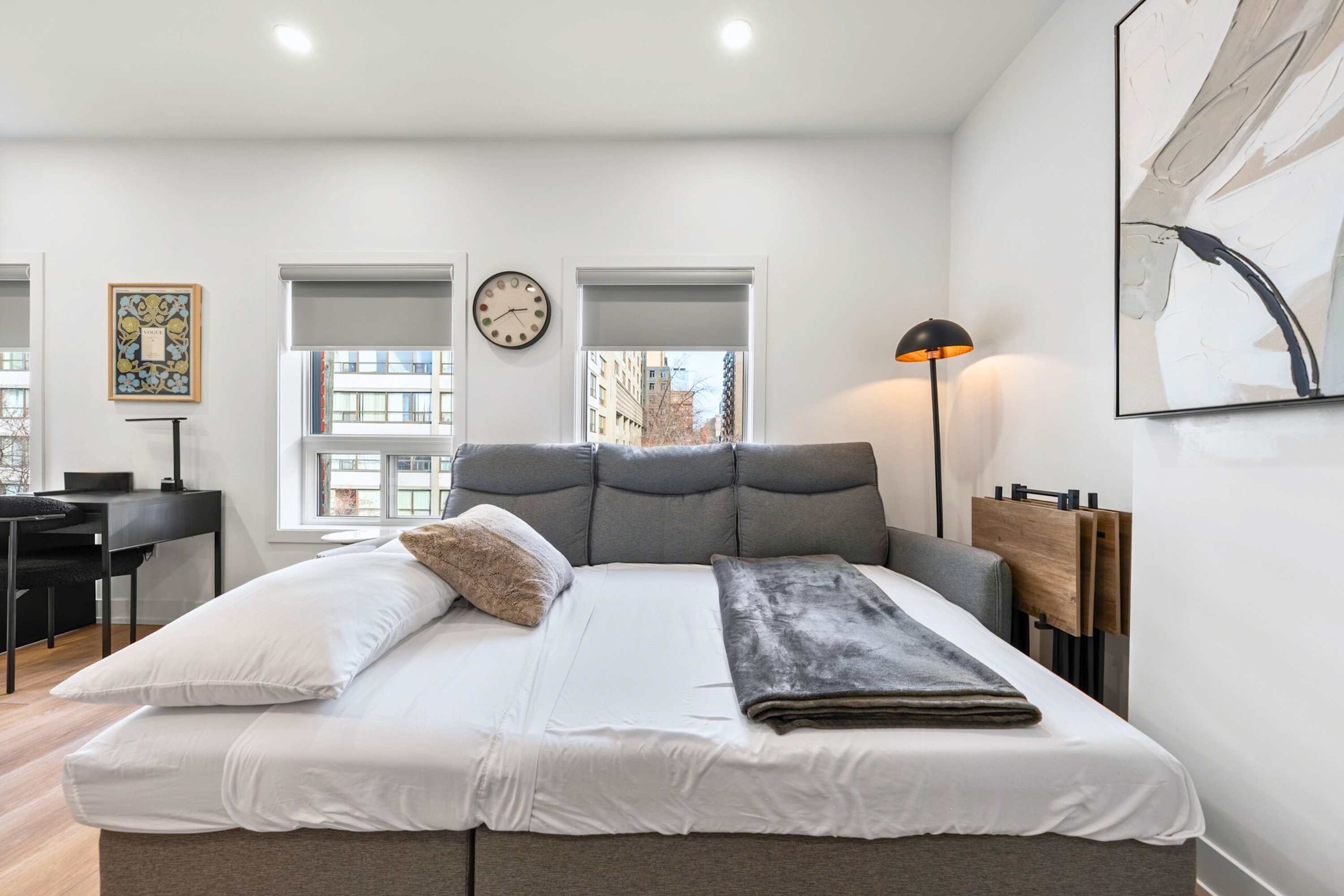 The gray sectional sofa in the living room is converted into a comfortable bed with white sheets and a gray blanket, highlighting its dual function as a sofa bed.