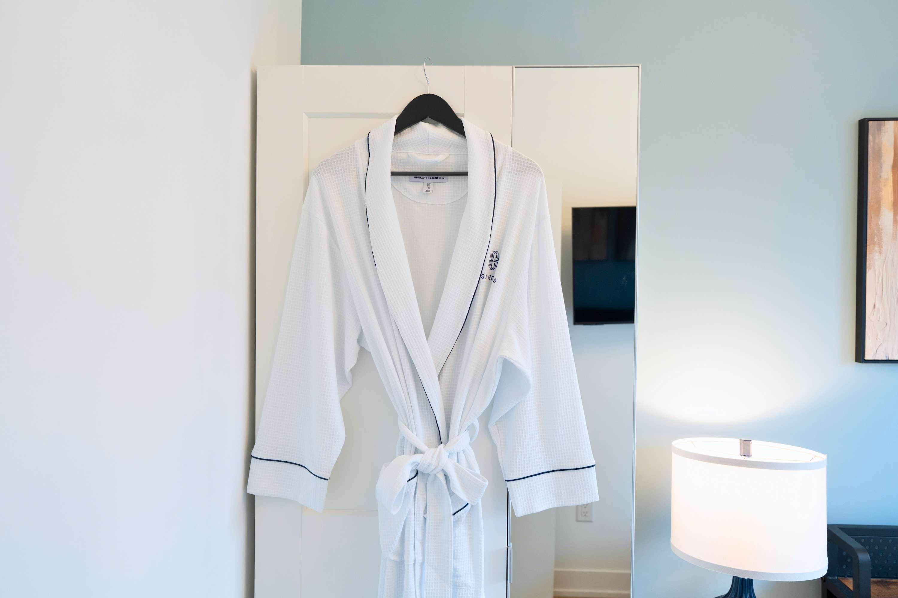 A white waffle-knit bathrobe with dark piping and an embroidered logo hangs neatly on a hanger from a white door. A modern lamp and a mirror are visible in the background, suggesting a bedroom or dressing area.