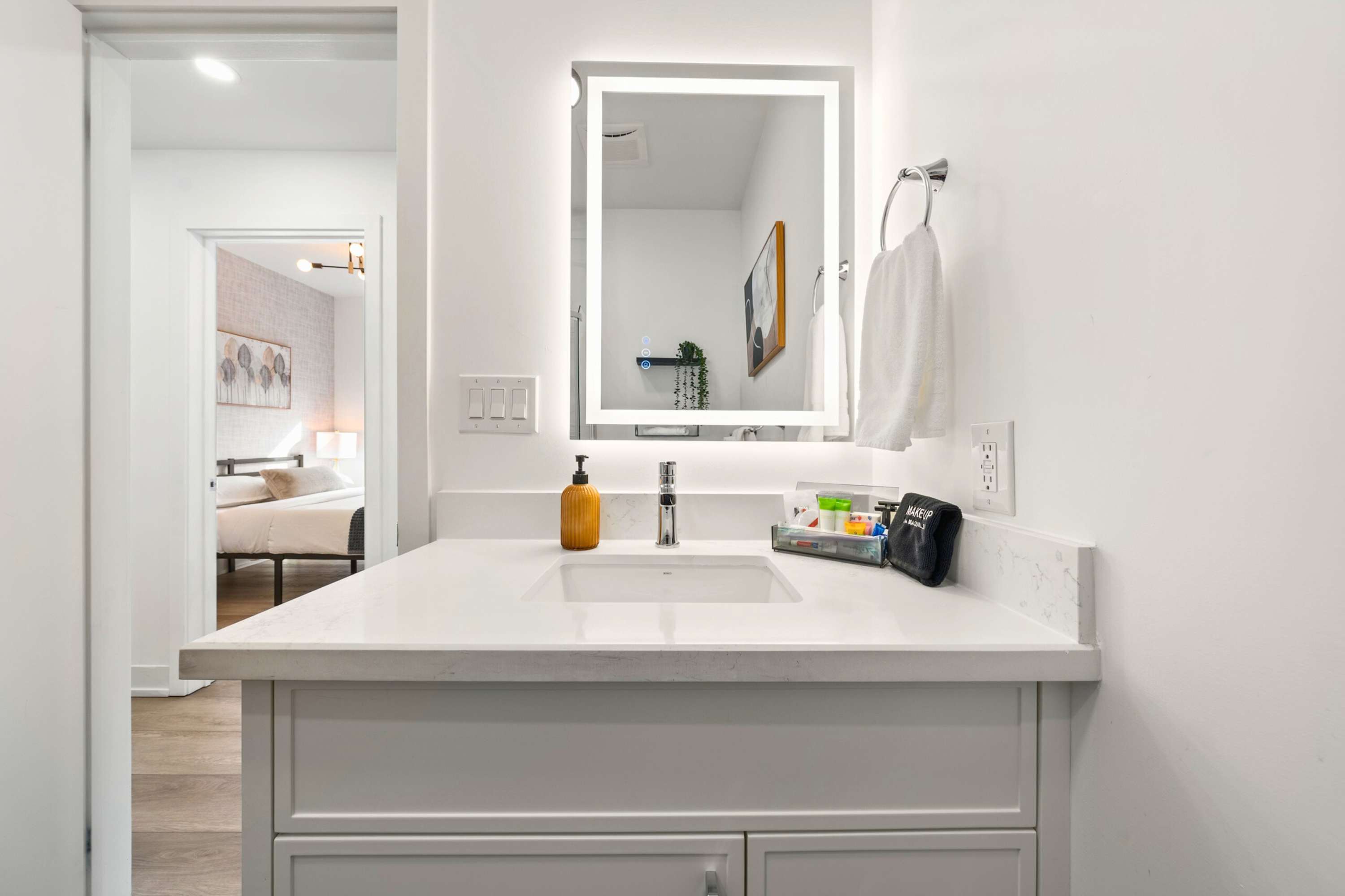 Modern bathroom vanity with a white countertop and integrated sink, featuring a large mirror with backlighting. A yellow soap dispenser and various toiletries are neatly arranged on the counter, with a bedroom visible through the open doorway.