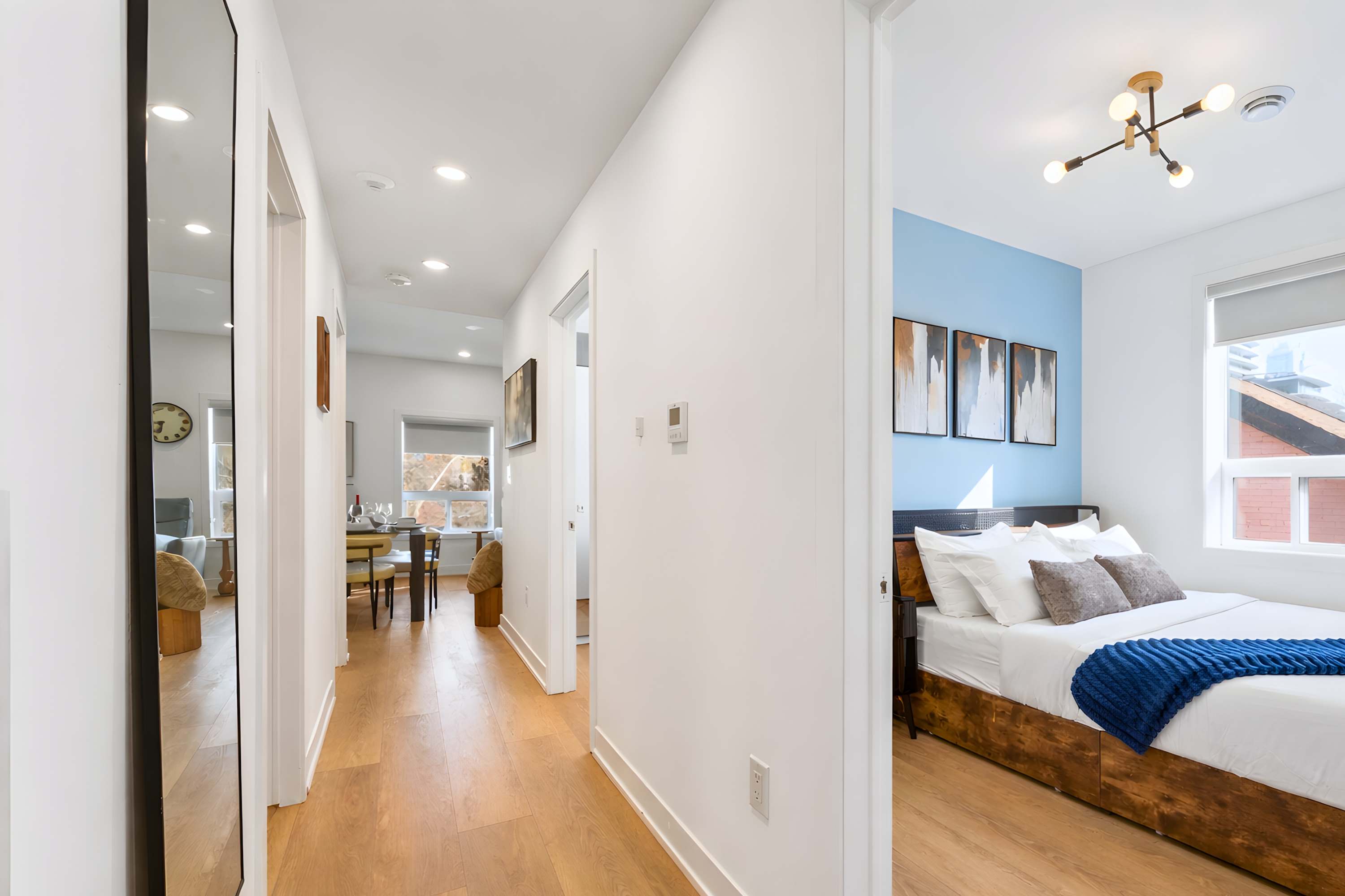 Bright, minimalist hallway with light wood flooring and white walls, featuring recessed lighting and several doorways leading to other rooms. A full-length mirror stands on the left, reflecting a dining area in the distance.