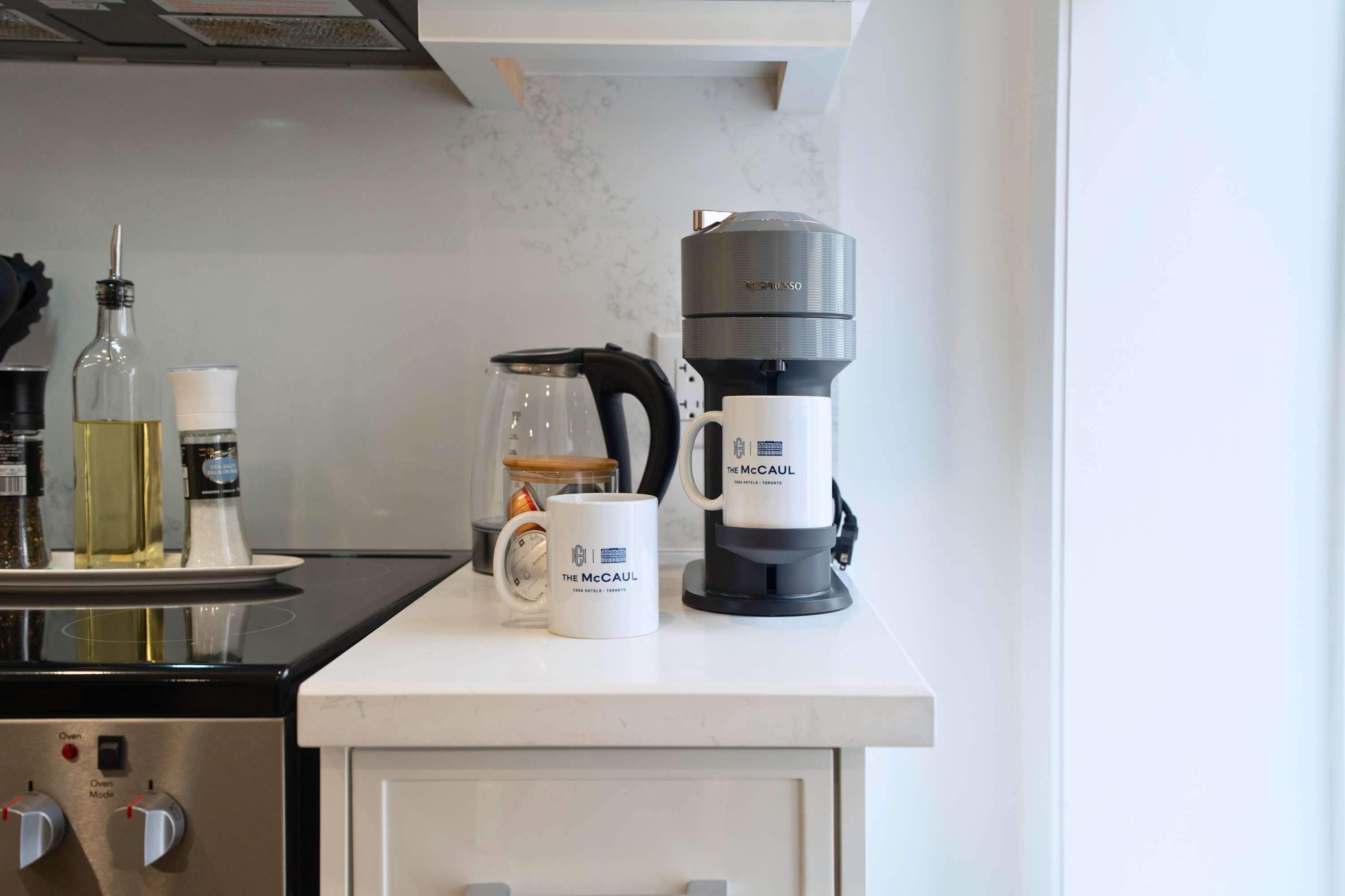 Close-up view of a kitchen countertop featuring a Nespresso coffee machine, an electric kettle, and two white mugs with "The McCaul" logo, alongside olive oil and spice grinders.