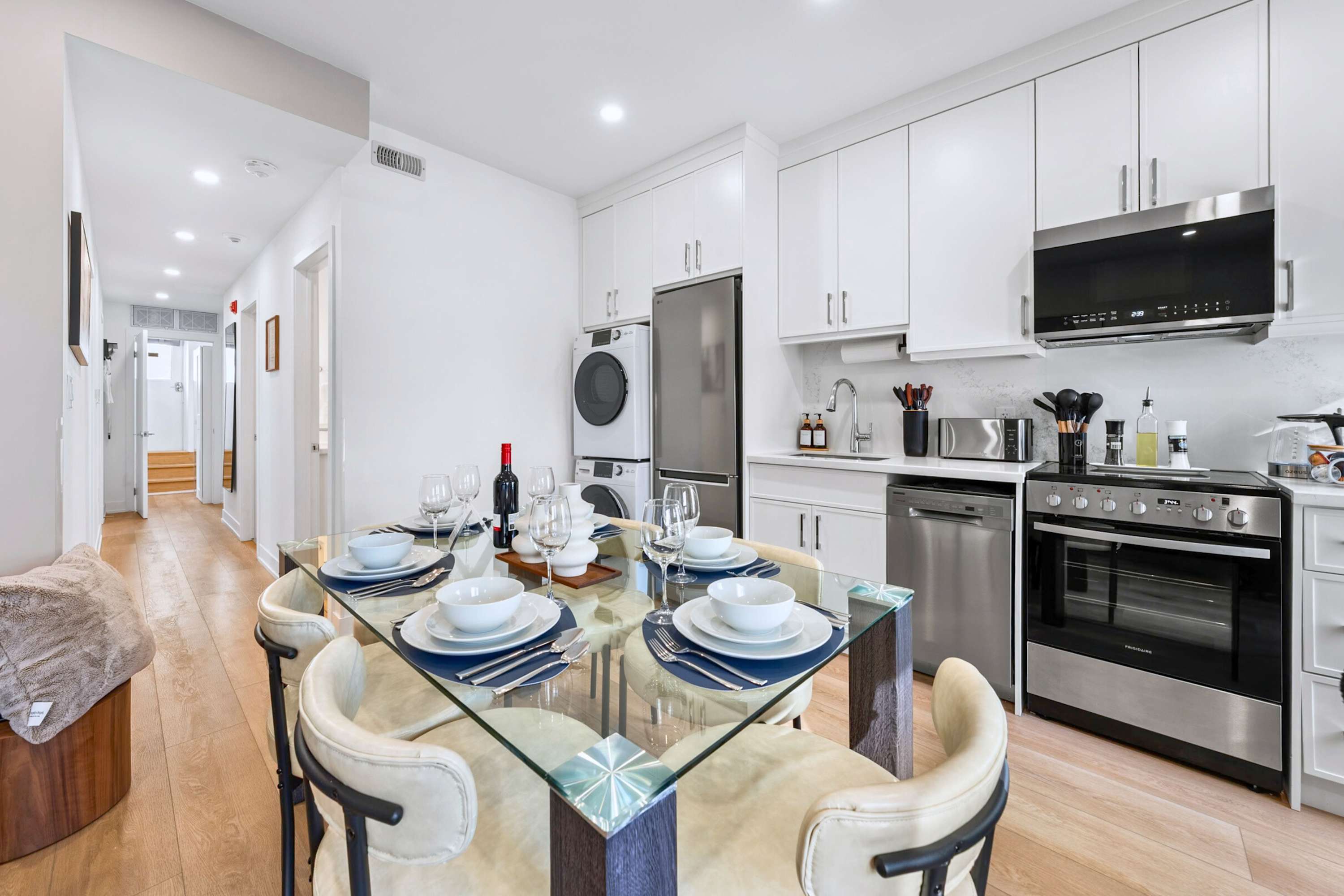 View of the open-concept kitchen and dining area, showing the glass-top dining table set for six, and the modern kitchen with white cabinets and stainless steel appliances. A hallway is visible to the left.