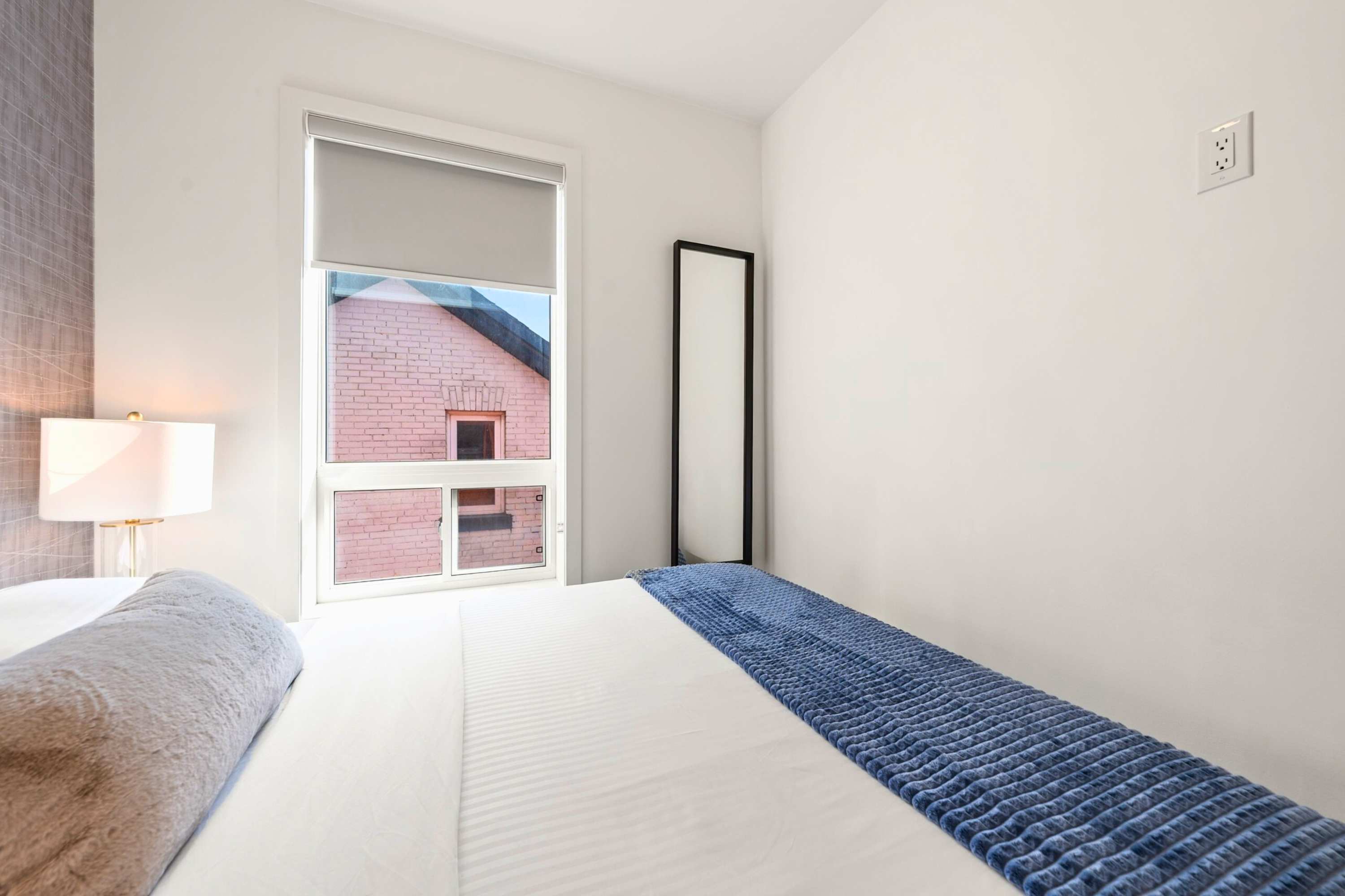 View of bedroom-1 showing a full-length mirror leaning against a white wall, a large window with a roller shade, and a bed with a blue throw blanket. The window offers a view of a brick building.