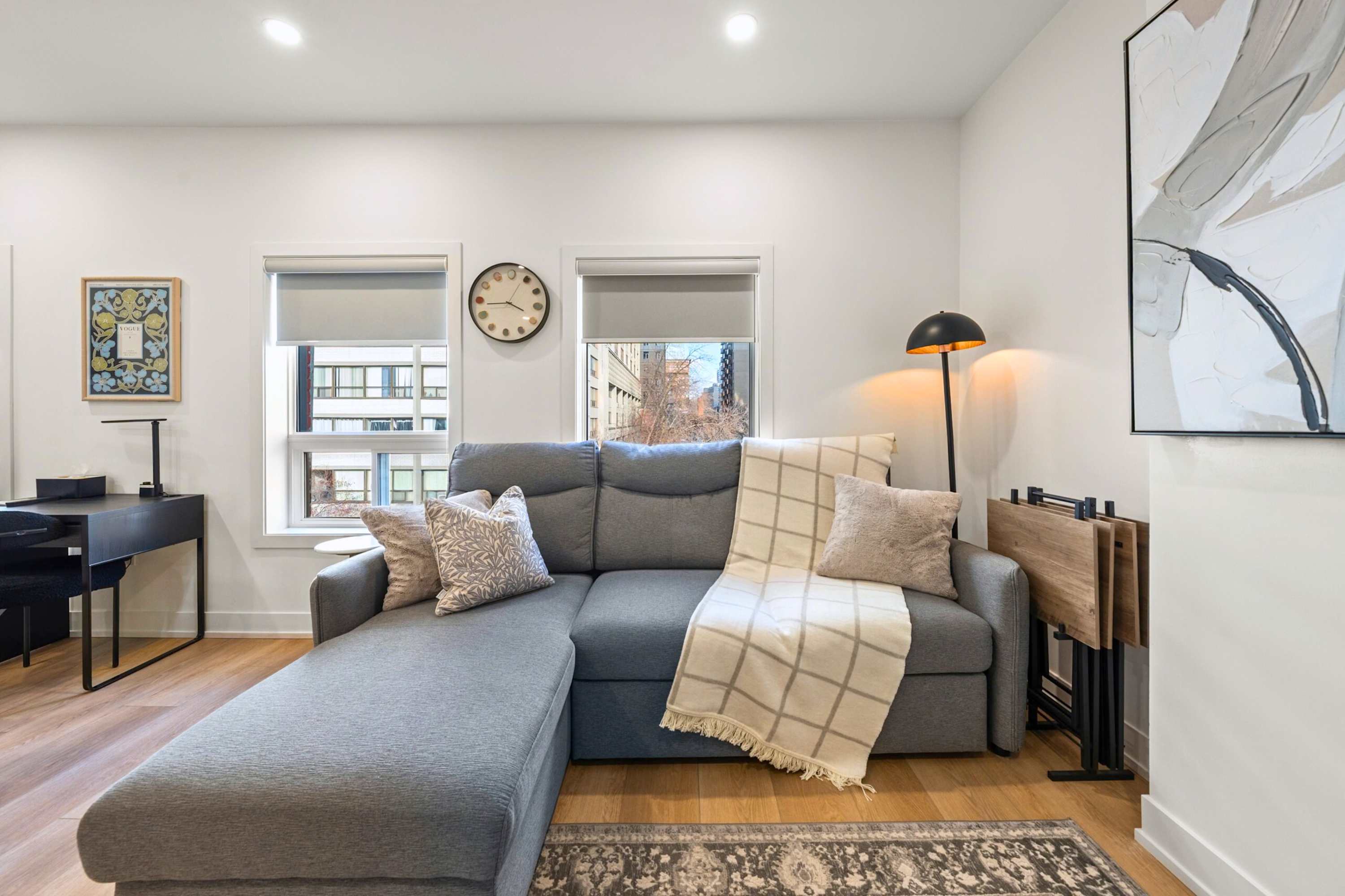Living room with a gray sectional sofa, two large windows with blinds, a round clock on the wall, and a black desk in the corner. A floor lamp and a set of folding tables are also visible.