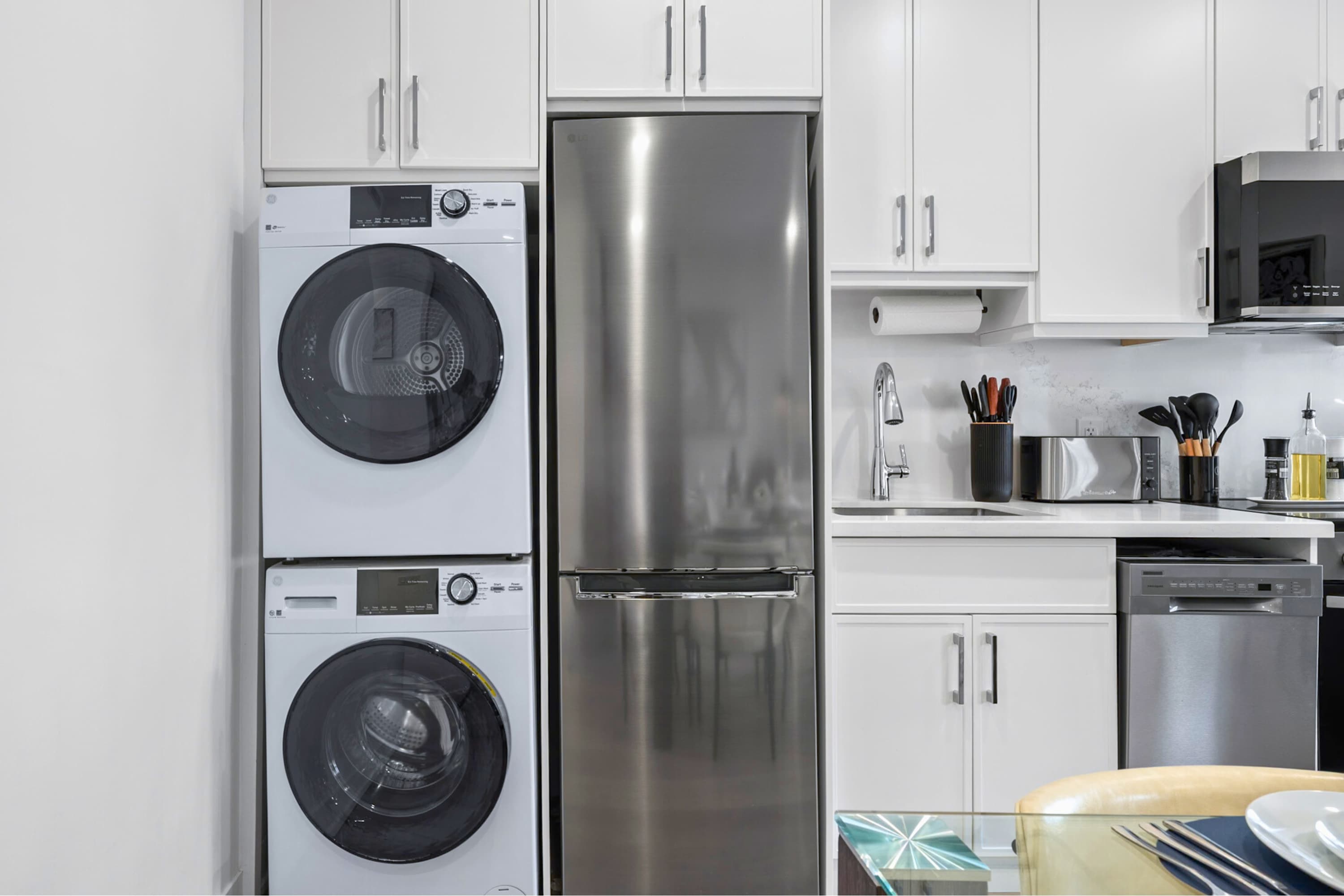 Stacked white front-loading washer and dryer units are neatly integrated into the kitchen area, positioned next to a stainless steel refrigerator and white cabinetry.