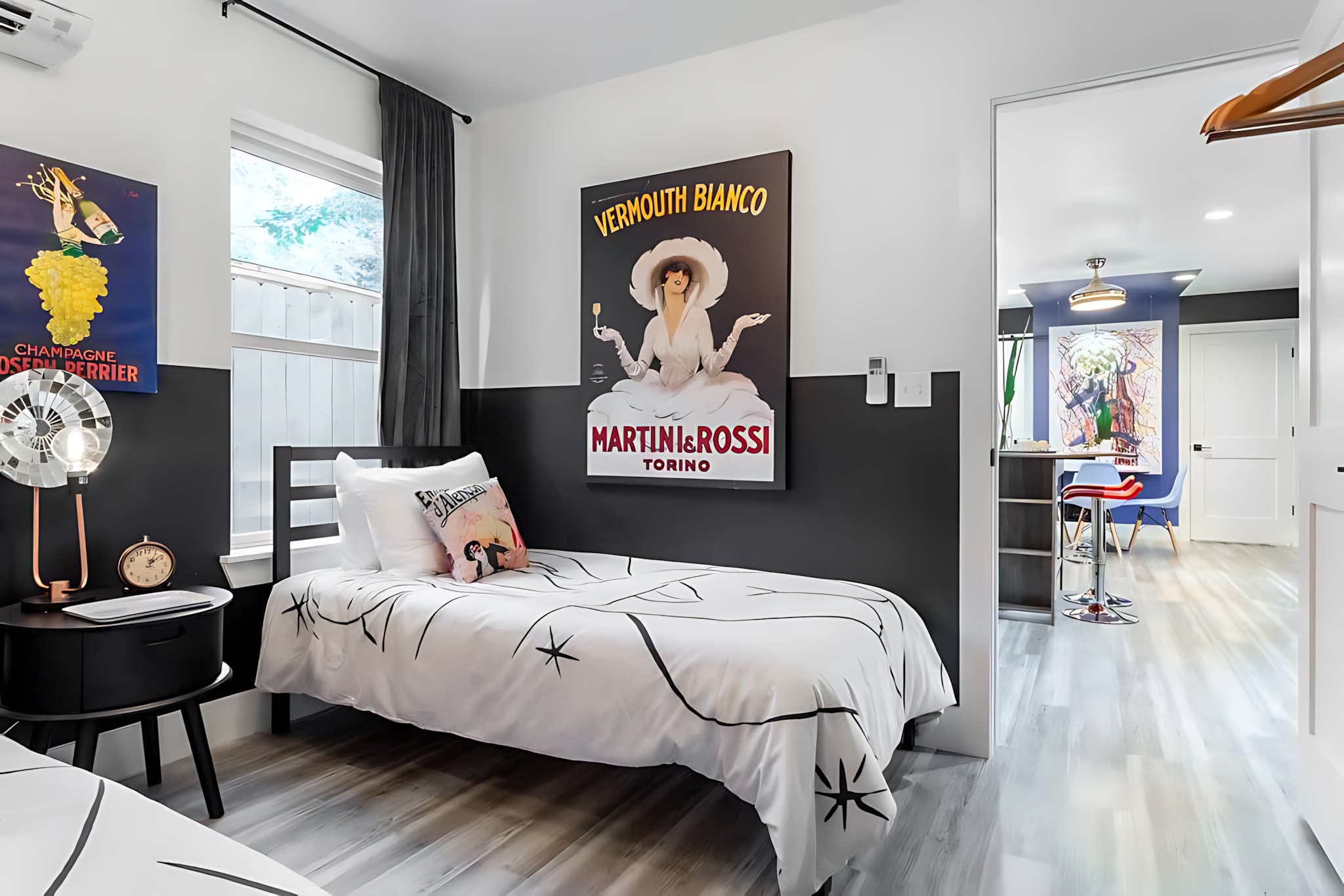 A stylish bedroom with a black and white color scheme, featuring two twin beds with graphic bedding, and vintage-style advertising posters on the walls. A window is covered by dark curtains, and a modern lamp sits on a black nightstand.