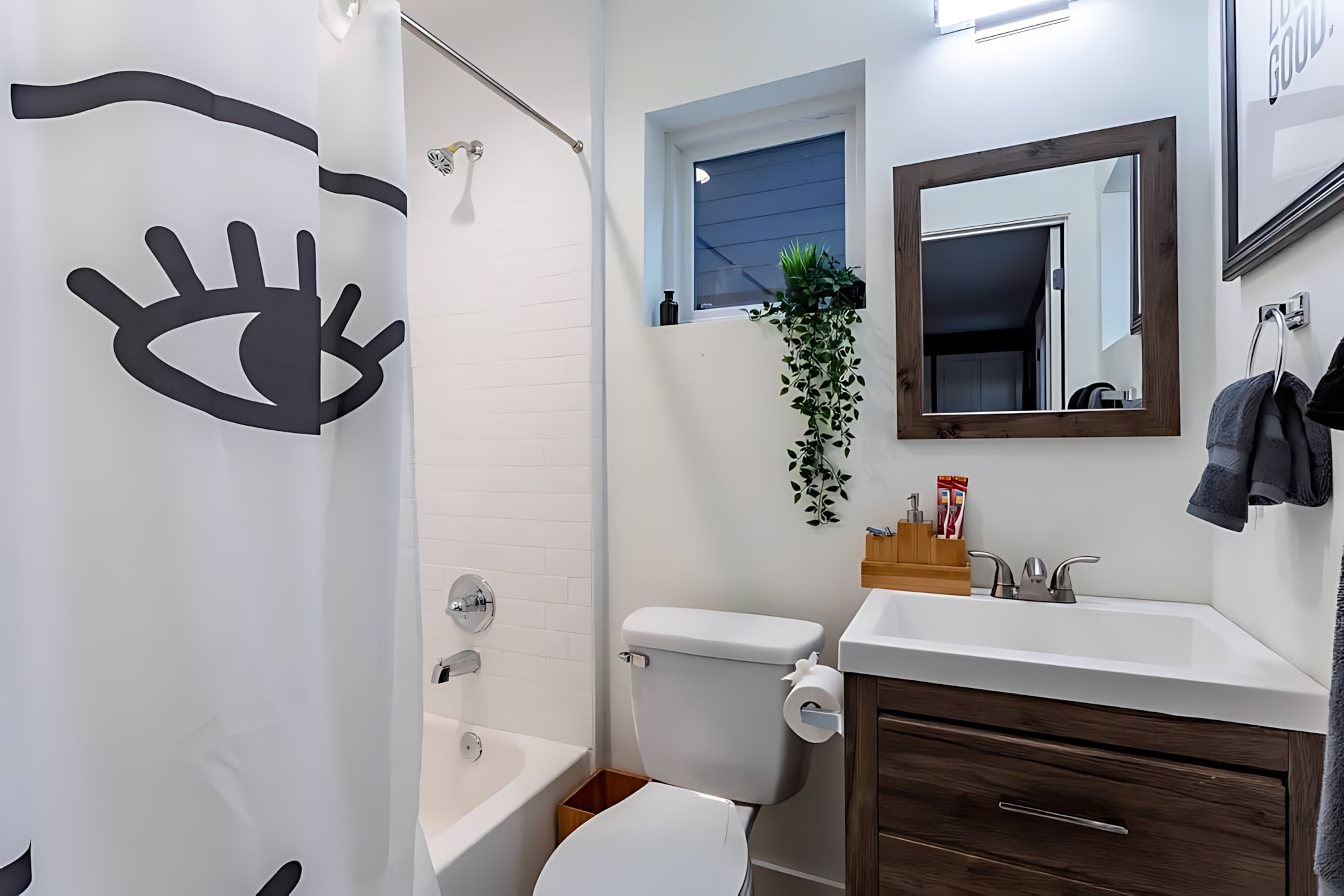 A bright bathroom with white subway tile, a white tub/shower combo featuring a black and white eye-patterned curtain, and a dark wood vanity with a white sink. A small window is positioned above the toilet.