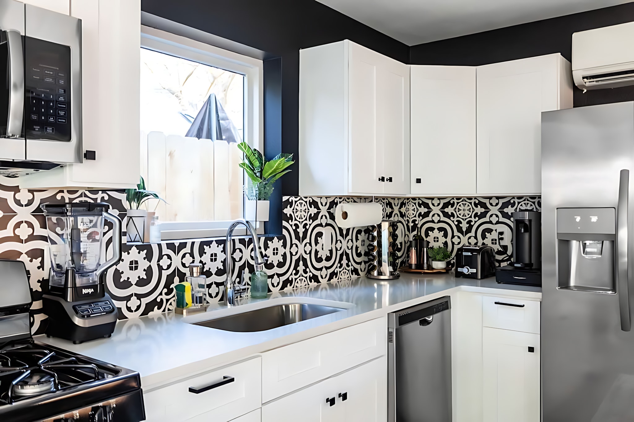 View of the kitchen sink area with a stainless steel sink, modern faucet, and a window above. The black and white patterned tile backsplash continues around the corner, with a paper towel holder and various kitchen appliances visible.