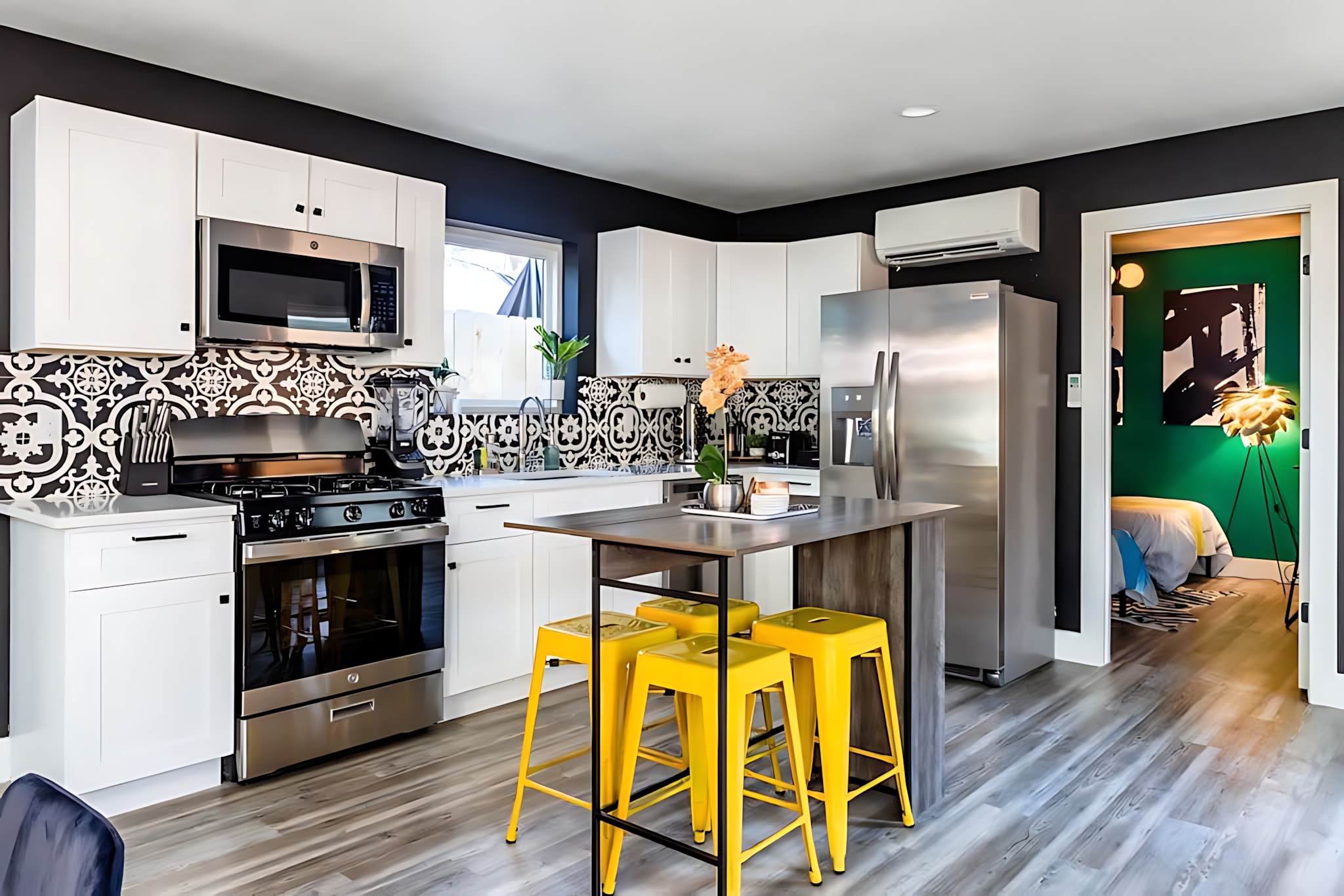 Wide shot of a modern kitchen with white cabinets, stainless steel appliances, and a black and white patterned tile backsplash. A dark wooden island with three bright yellow bar stools provides a dining or casual seating area.