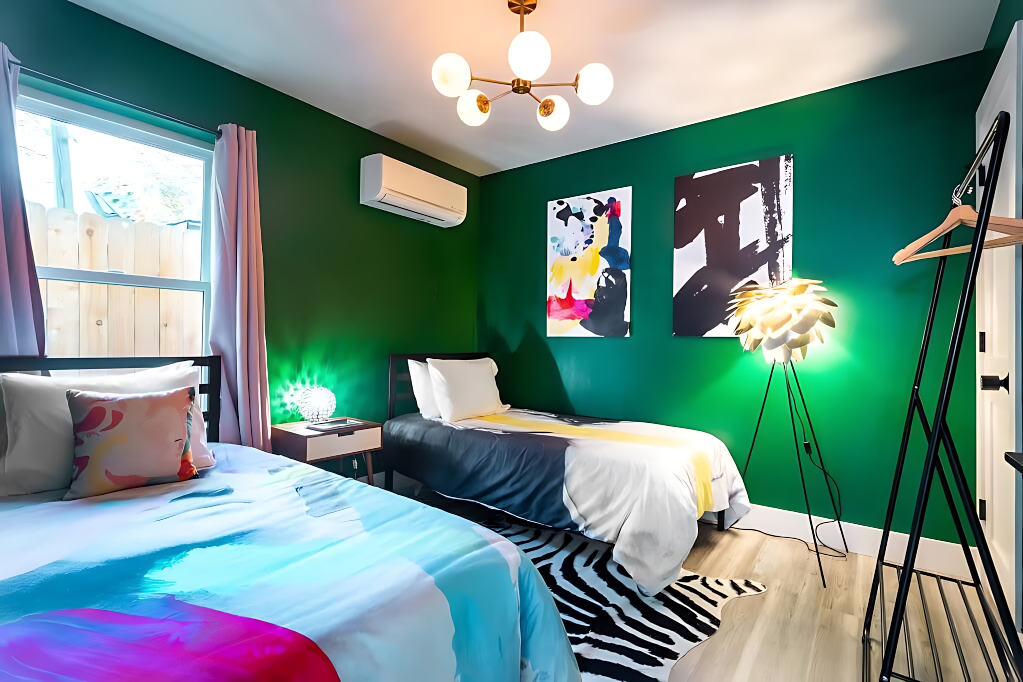 A colorful bedroom with dark green walls, two twin beds featuring abstract bedding, and modern pop art on the walls. A large window is covered by light pink curtains, and a unique floor lamp adds character.