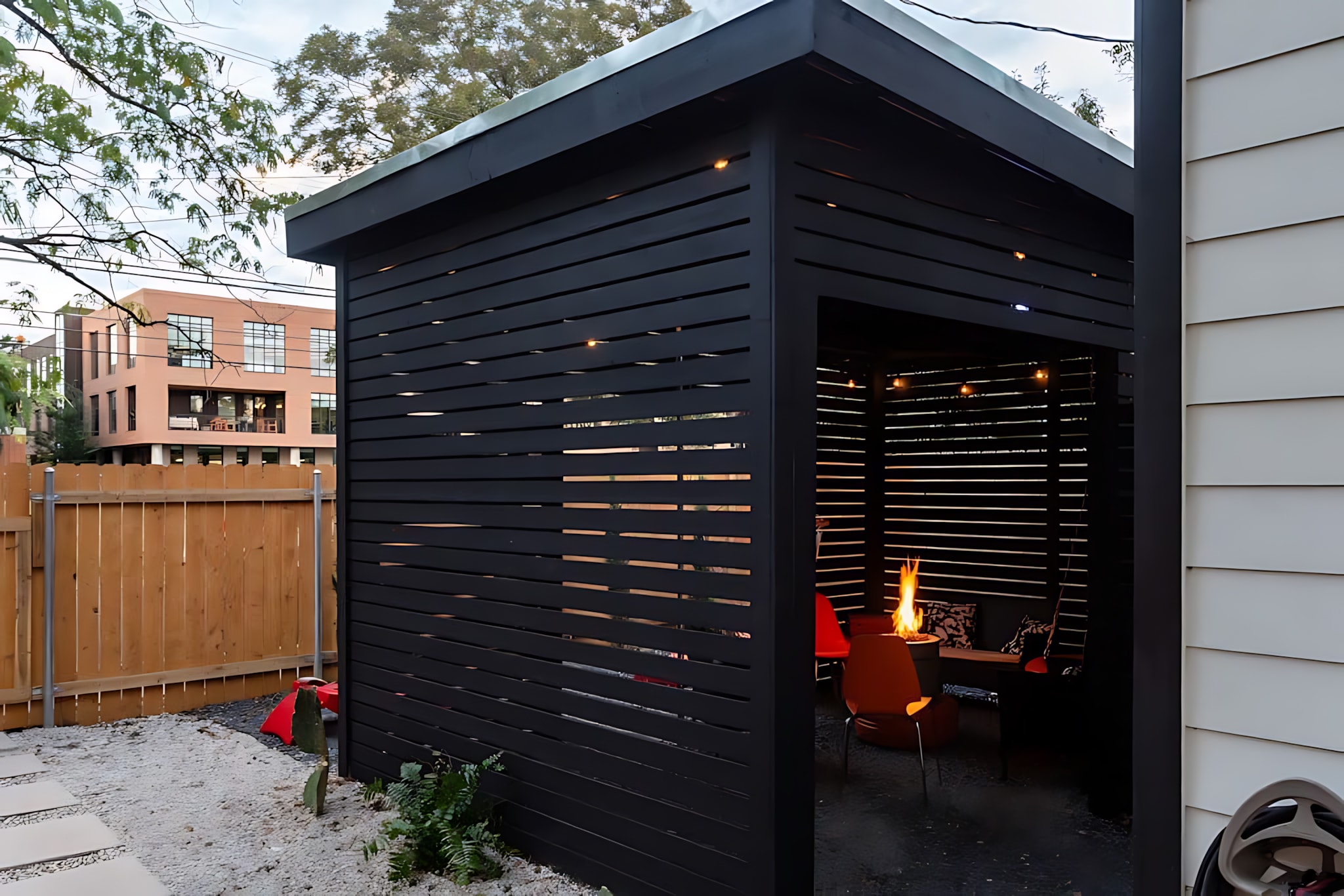 The exterior of a modern black slatted outdoor structure, featuring a fire pit visible inside, stands next to a wooden fence and a gravel yard with stepping stones.