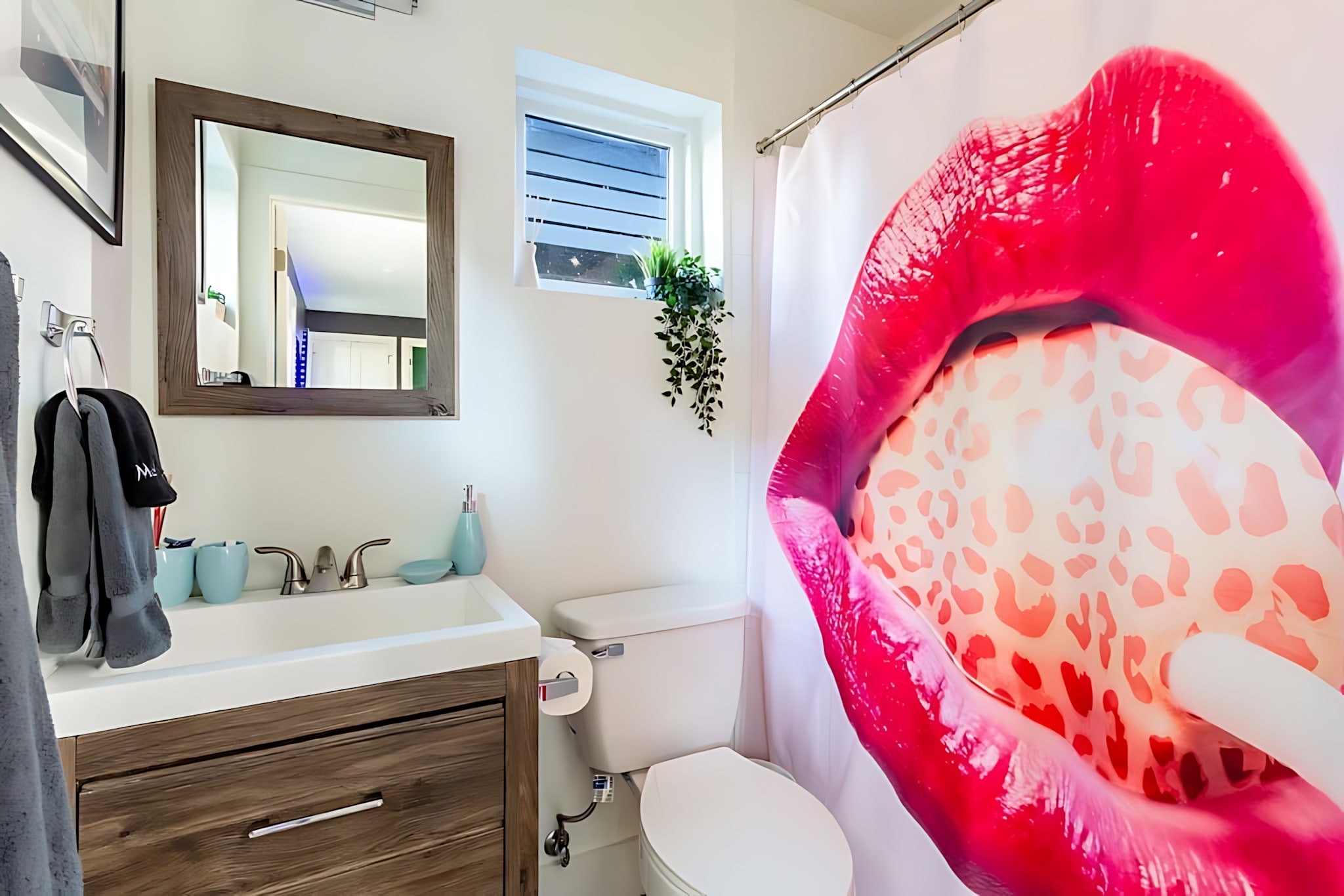 A vibrant bathroom with a white tub/shower combo featuring a bold red lips and leopard print shower curtain, a dark wood vanity with a white sink, and a small window above the toilet. White subway tiles line the shower area.