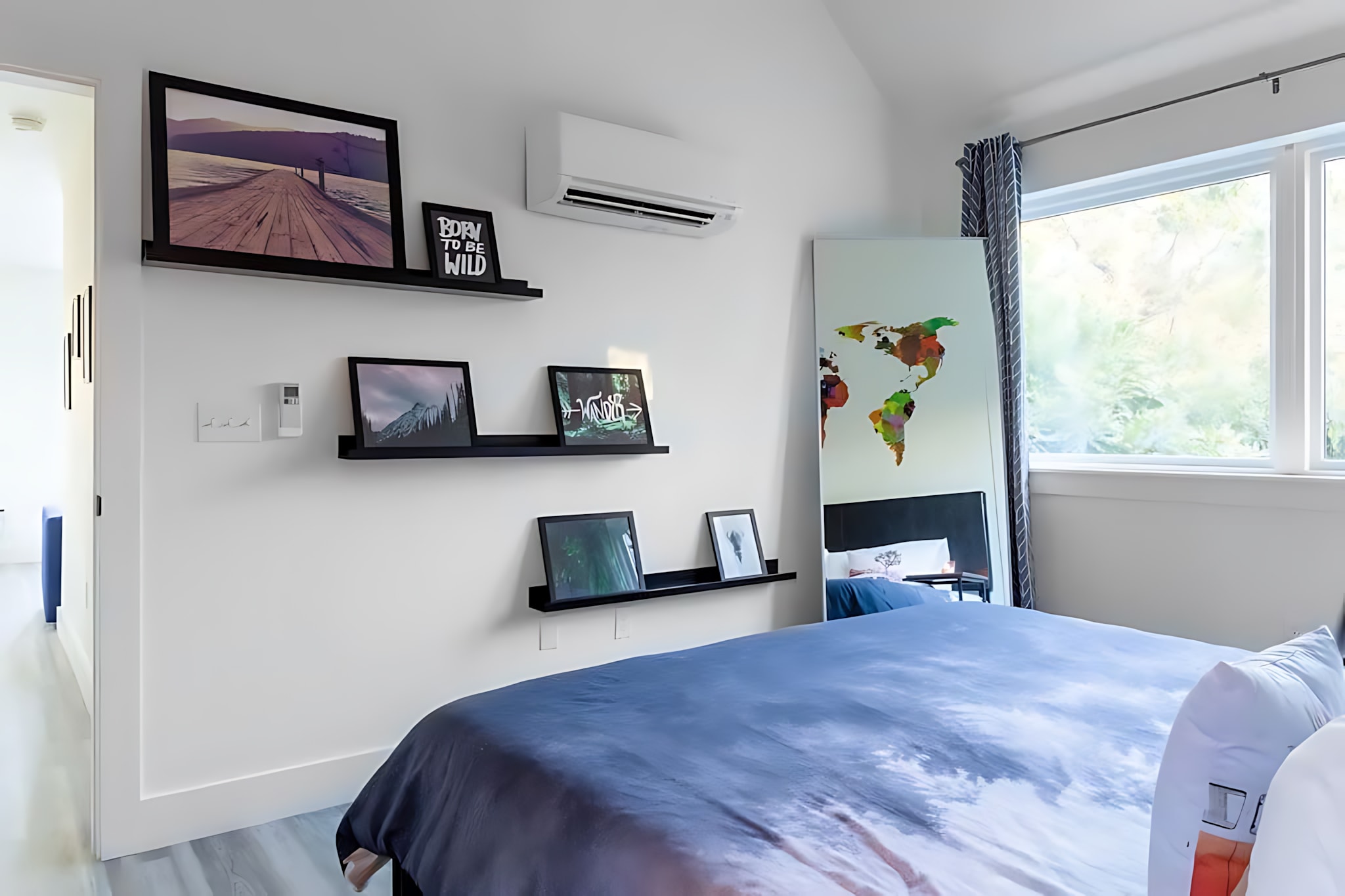 A bright bedroom with a bed covered in a blue and white patterned duvet, featuring black floating shelves displaying framed art, and a large window overlooking greenery.