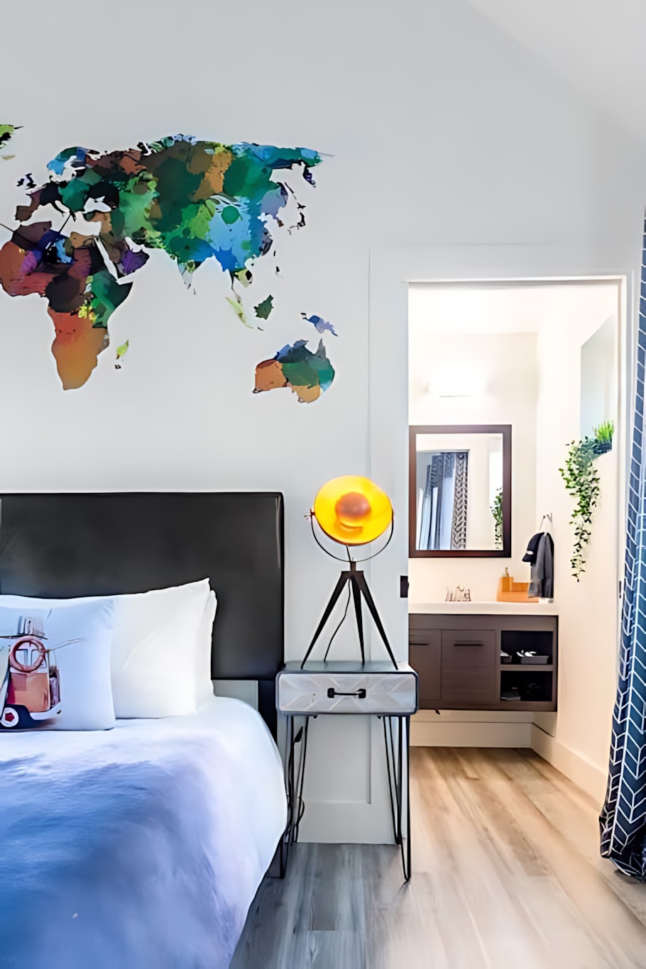 A modern bedroom with a black headboard and a bed covered in a blue duvet, featuring a colorful world map decal on the wall and a unique tripod lamp on a bedside table. An en-suite bathroom is visible through the doorway.