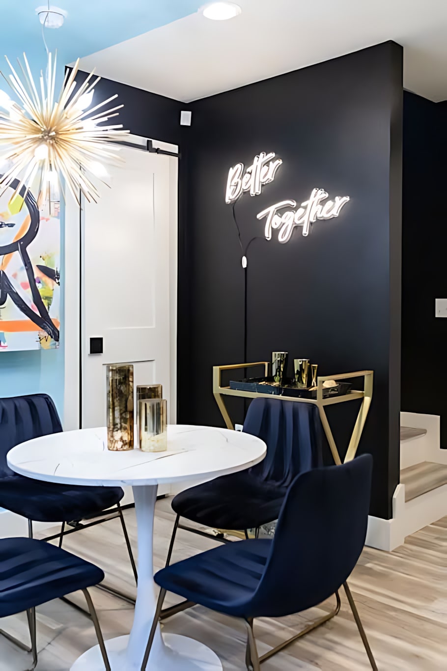 A cozy dining nook with a round white table and four dark blue velvet chairs. A 'Better Together' neon sign illuminates a dark accent wall, and a gold bar cart with glassware is visible in the corner.