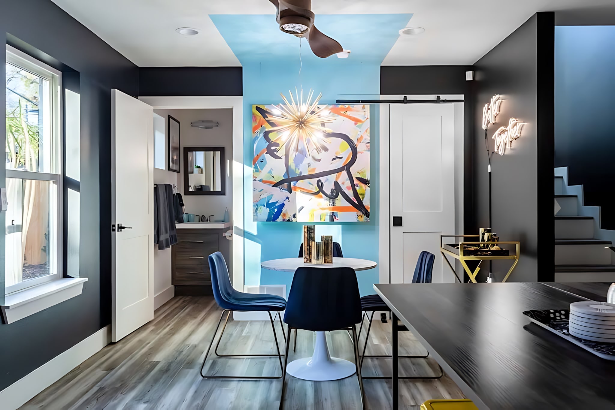 Bright dining area with a white round table and blue chairs, illuminated by a sputnik chandelier, set against a blue accent wall with abstract art and a blue ceiling panel.
