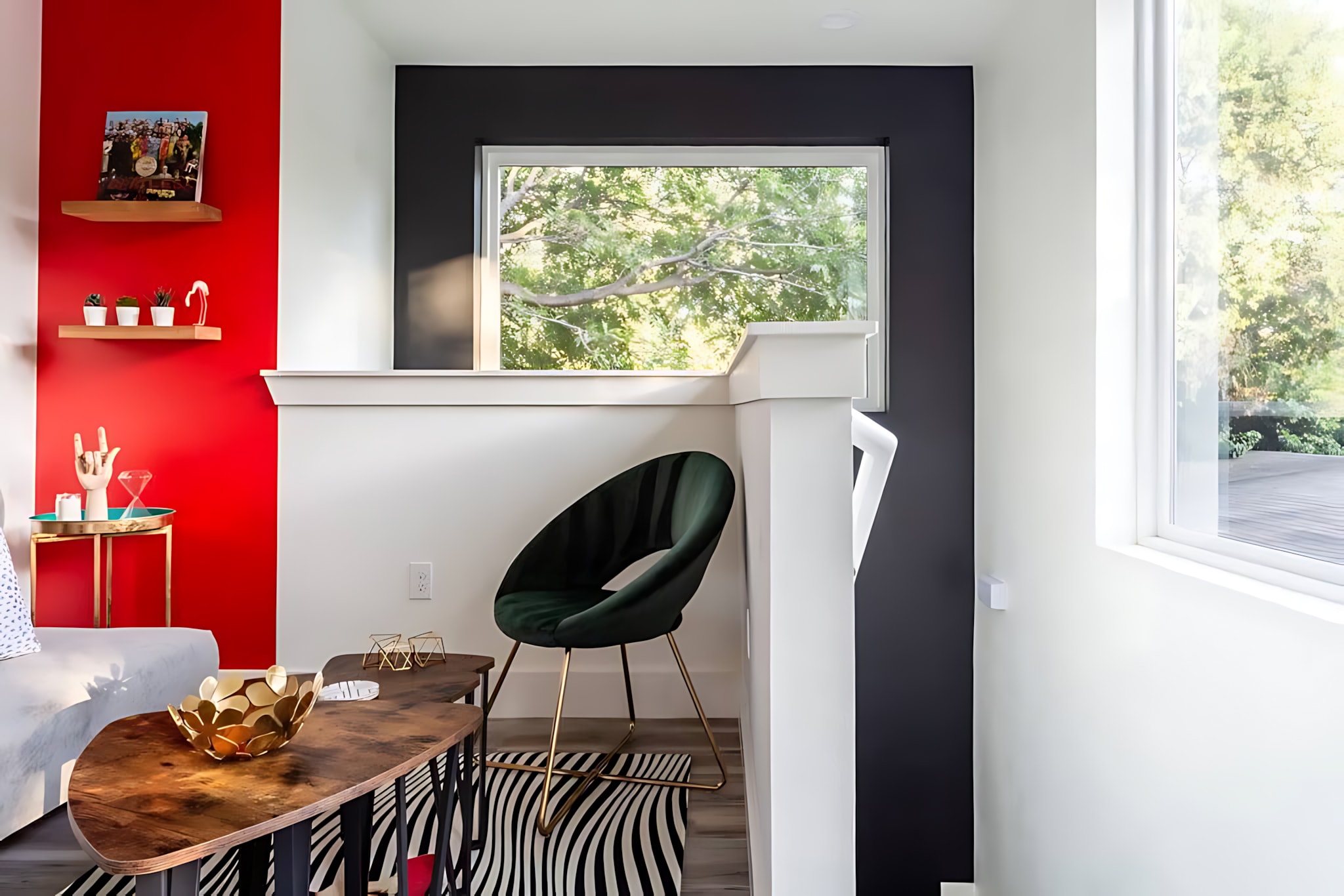 A bright and eclectic living space featuring a dark green velvet accent chair with gold legs on a black and white striped rug. A multi-level wooden coffee table is in the foreground, with a red accent wall and large windows providing natural light.