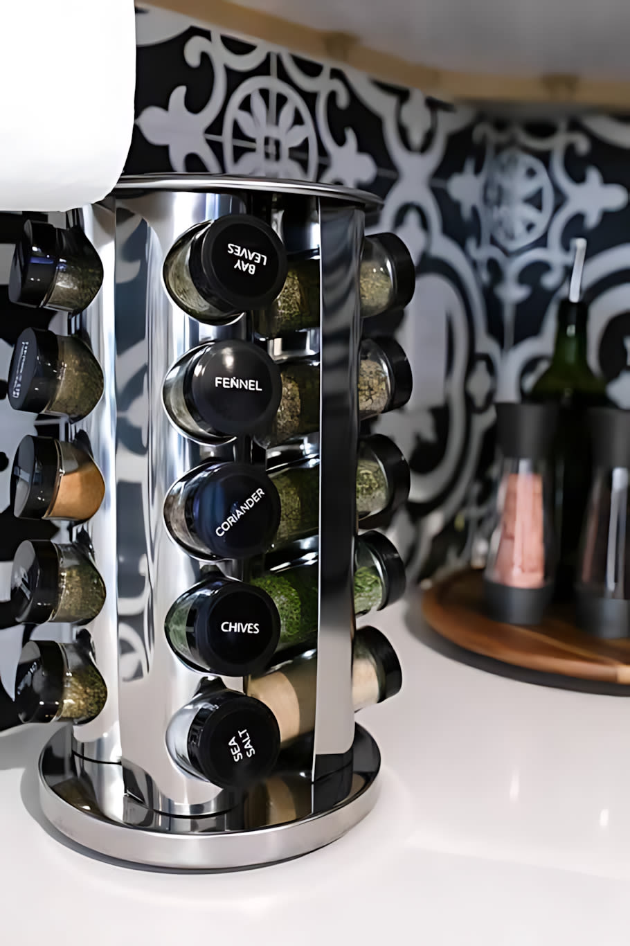 A chrome rotating spice rack filled with labeled spice jars, including 'BAY LEAVES', 'FENNEL', 'CORIANDER', 'CHIVES', and 'SEA SALT'. The black and white patterned tile backsplash is visible behind it.