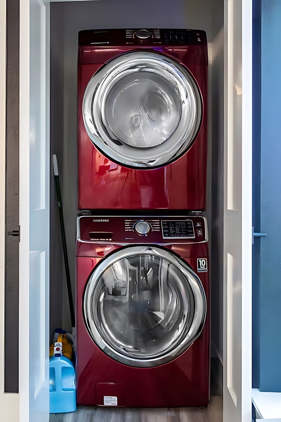 A vibrant laundry closet featuring a red stacked front-loading washer and dryer unit, with cleaning supplies visible on the floor. The appliances are housed within a narrow closet with white doors.