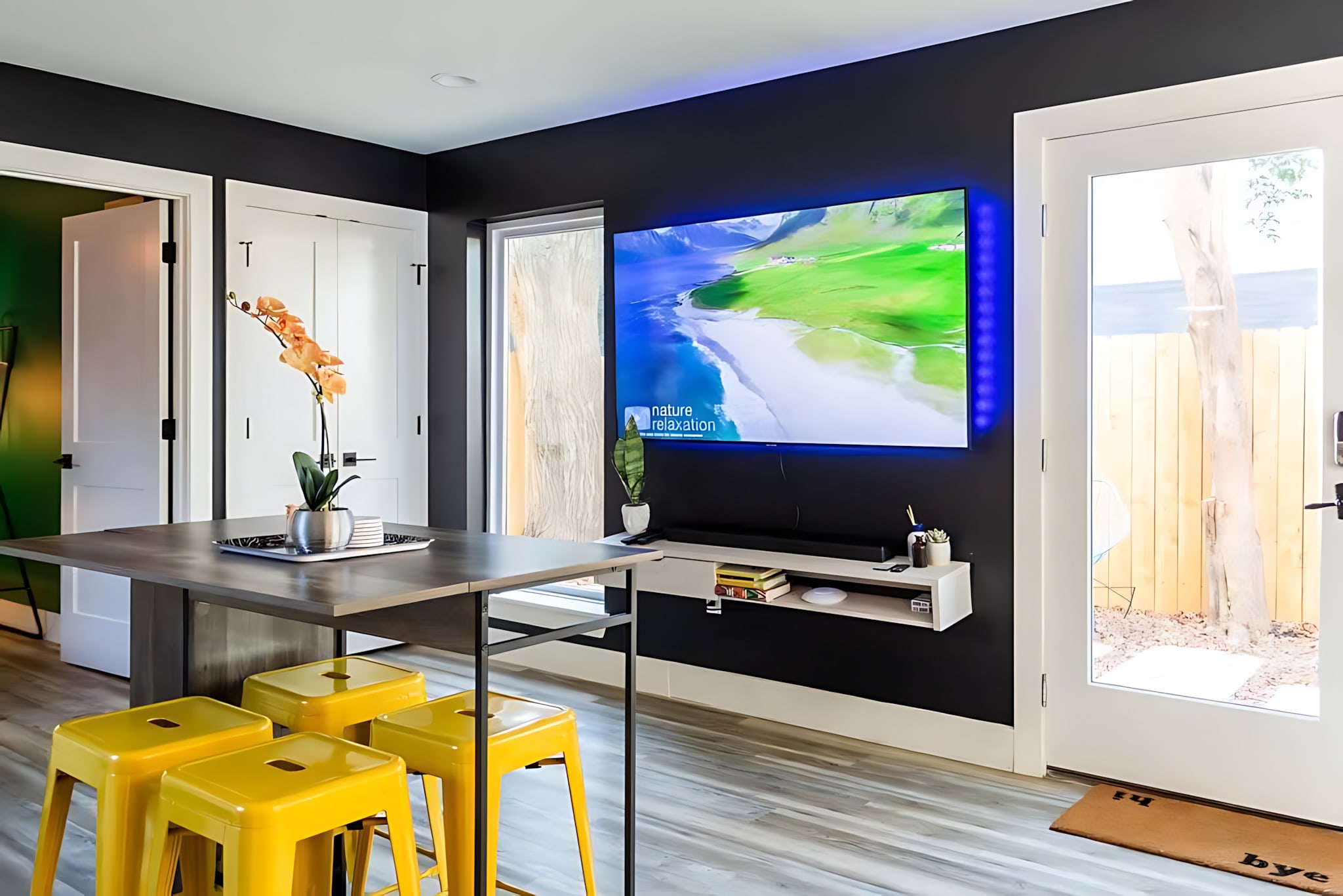 A bright living space with a large wall-mounted TV displaying a nature scene, accented by blue backlighting against a dark wall. A wooden island with yellow bar stools is in the foreground, and a glass door leads to an outdoor area.