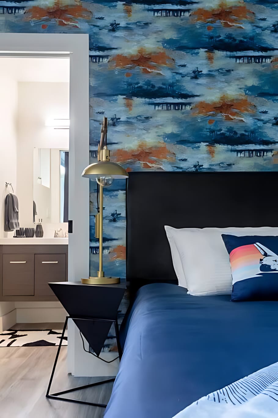 A close-up of a bedside in a bedroom, showing a black headboard, blue bedding, a decorative pillow, and a gold-accented lamp on a modern black bedside table. An en-suite bathroom is visible.