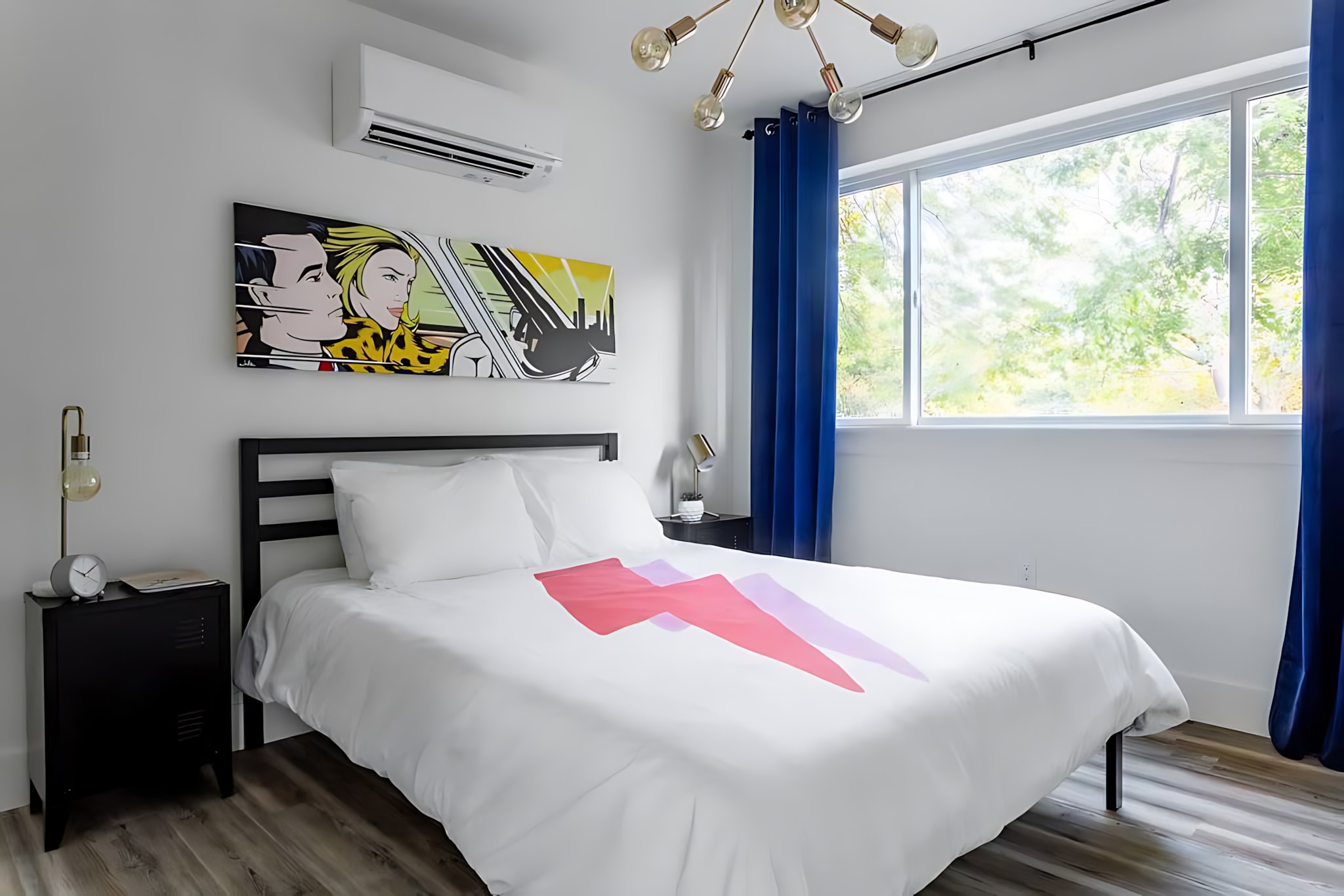 A modern bedroom featuring a queen-sized bed with a white duvet adorned with pink and purple lightning bolt graphics, a black metal headboard, and pop art on the wall. Blue curtains frame a large window, and an AC unit is mounted above the bed.